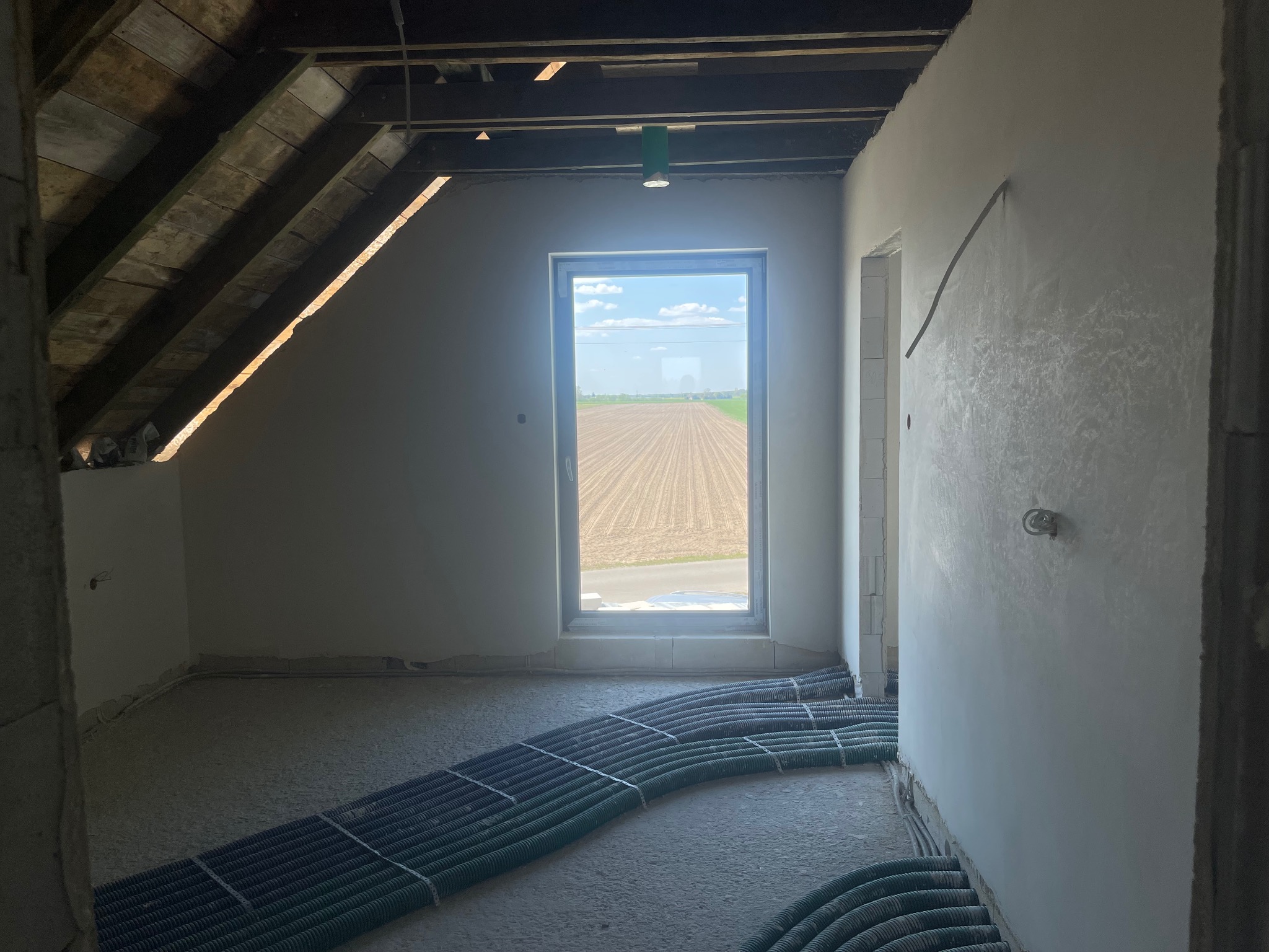 Poddasze w trakcie wykańczania: widoczne rury instalacyjne na podłodze, ściany po wstępnym wyrównaniu, surowy drewniany strop, okno z widokiem na pole.