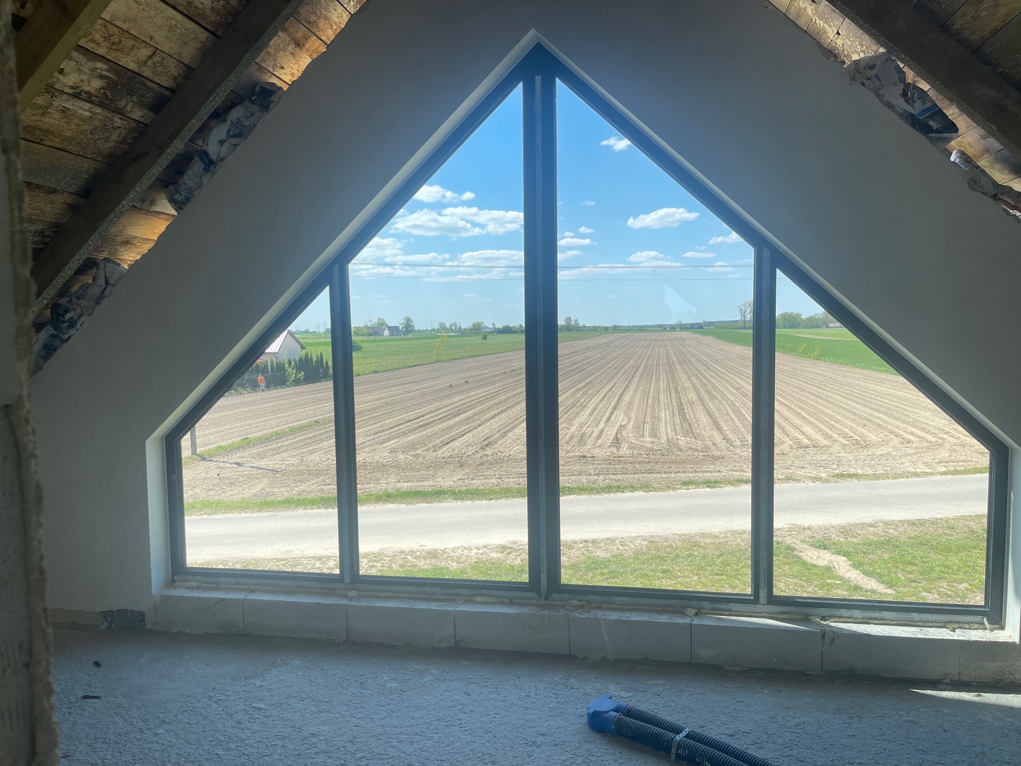 Poddasze w trakcie wykańczania z trójkątnym oknem z widokiem na pole, ściany przygotowane do dalszych prac wykończeniowych, widoczne instalacje elektryczne.