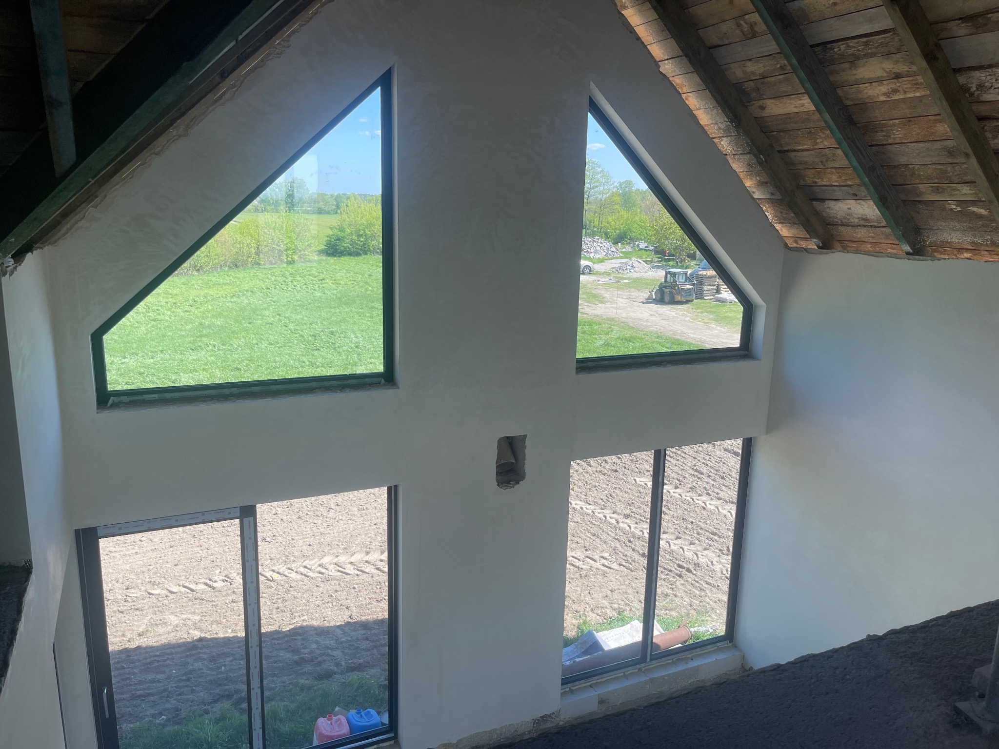 Wnętrze poddasza z surowym drewnianym stropem, ściany w trakcie wygładzania, duże okna z widokiem na pole i sprzęt budowlany.