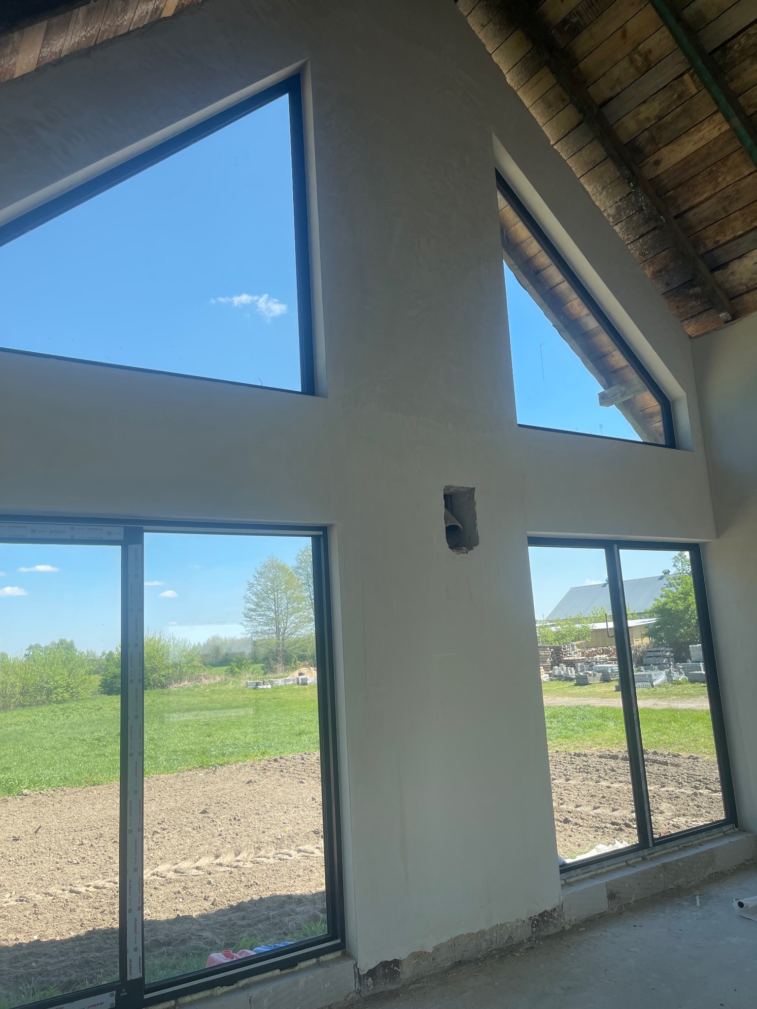 Wnętrze w stanie surowym, ściany po wstępnym wyrównaniu, widoczne okna z widokiem na zewnątrz, drewniana konstrukcja dachu.