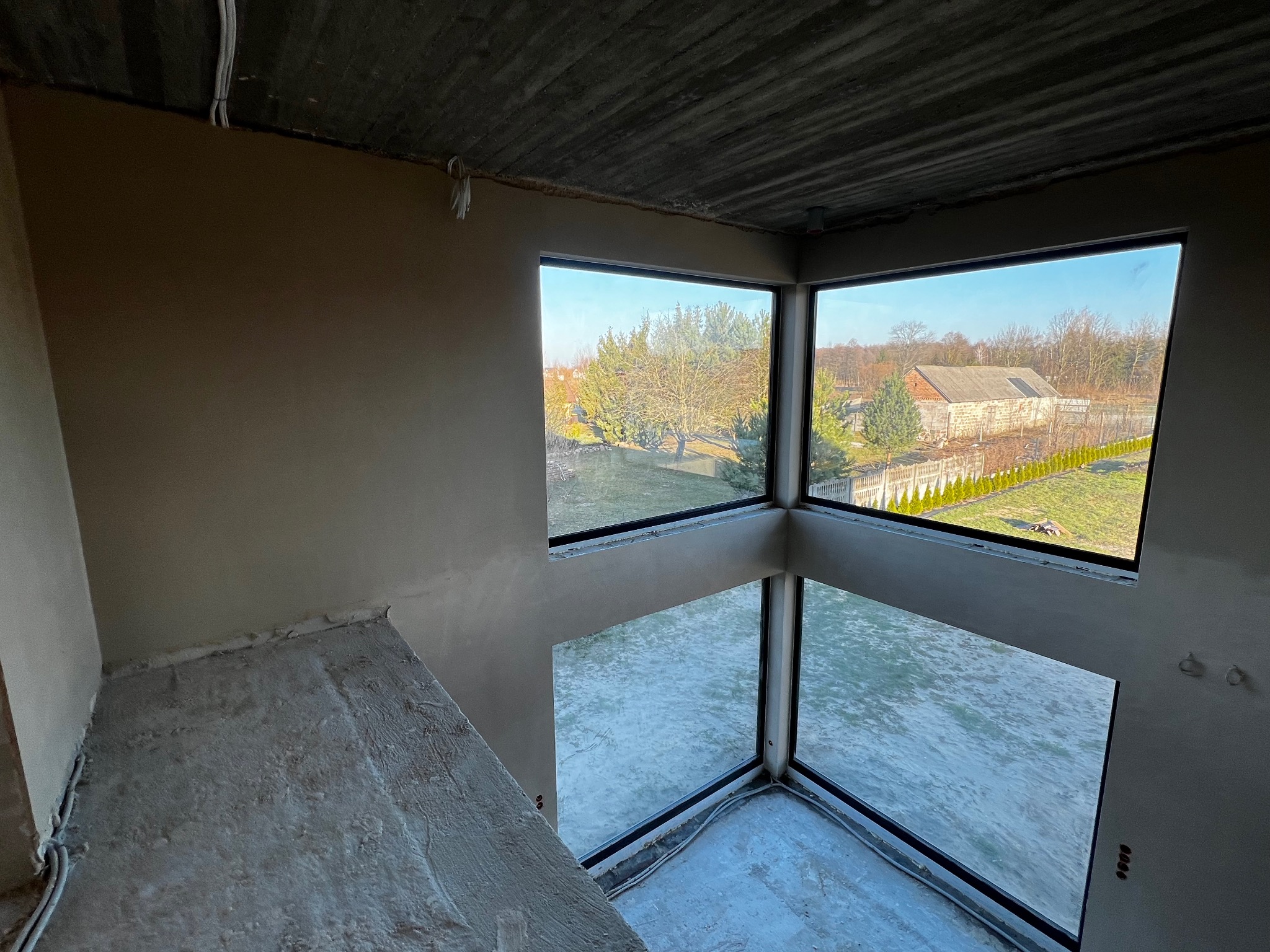 Surowy stan wnętrza z dużymi oknami narożnymi, ściany pokryte szarym tynkiem, widoczny surowy betonowy strop, instalacja elektryczna wyprowadzona ze ściany.