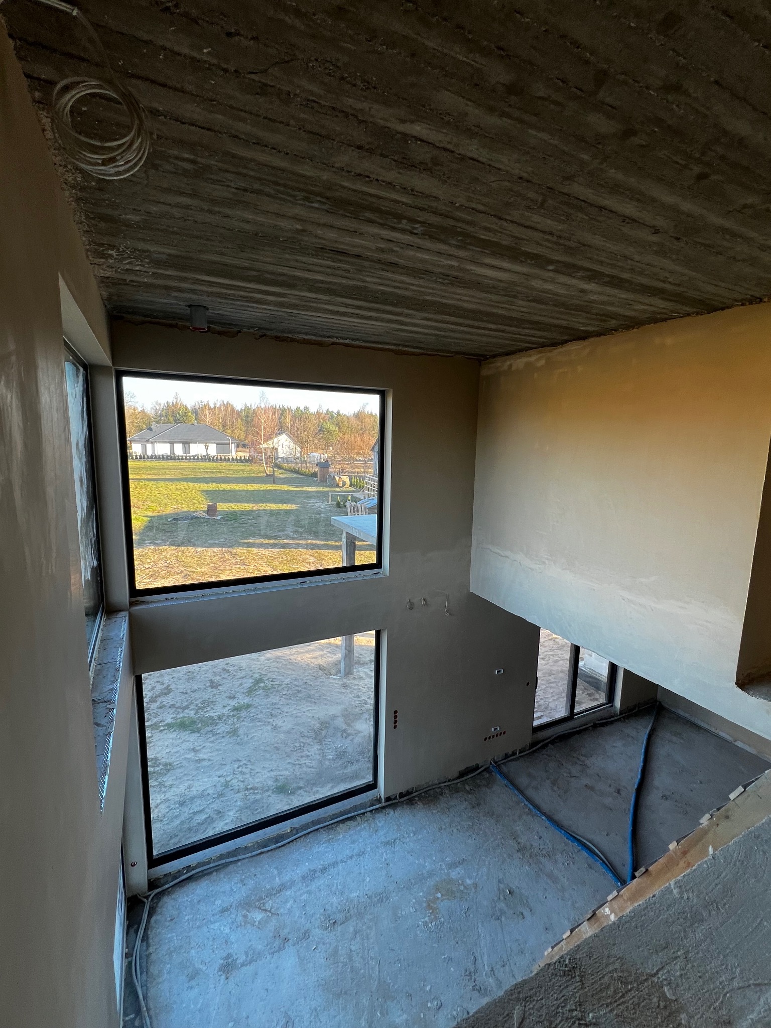 Surowy betonowy strop z widocznymi śladami szalunku w dwukondygnacyjnym wnętrzu w trakcie prac wykończeniowych, ściany pokryte tynkiem, duże okna z widokiem na zewnątrz.