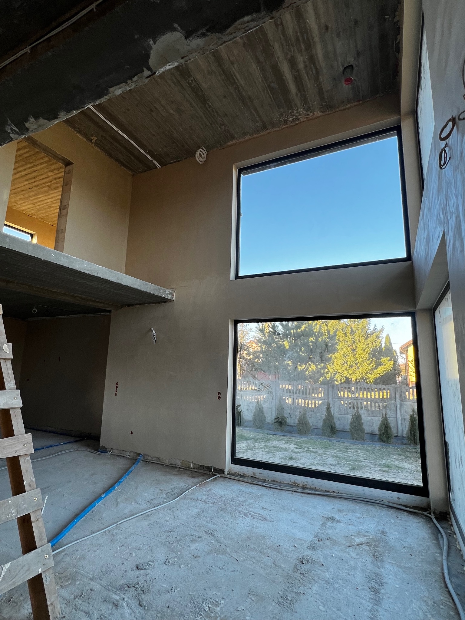 Wnętrze domu w trakcie prac wykończeniowych; ściany pokryte świeżym tynkiem, widoczne duże okna z widokiem na ogród, drewniana drabina i instalacje na podłodze.