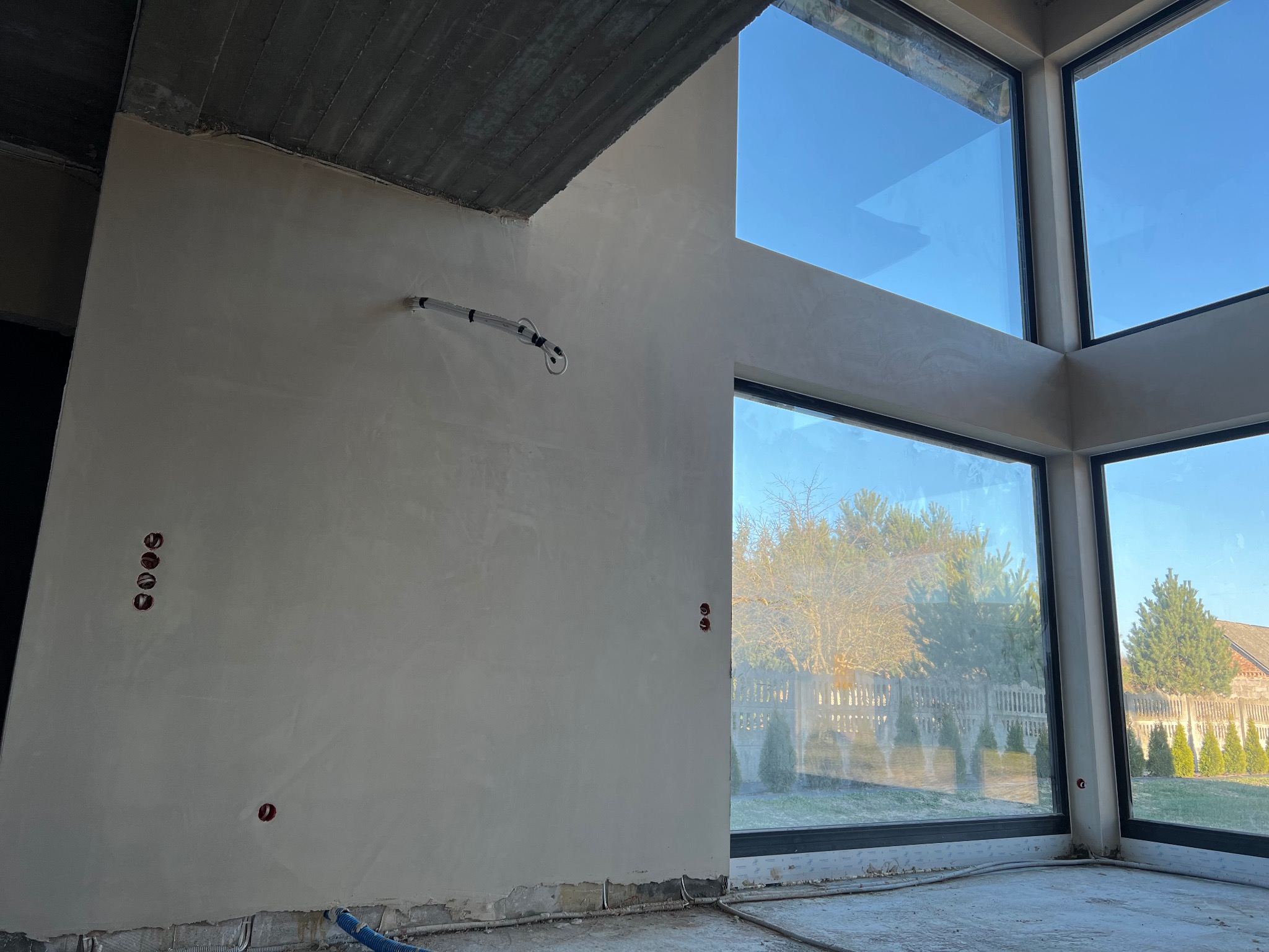 Ściana w surowym stanie po nałożeniu jasnej warstwy wykończeniowej, z widocznymi puszkami elektrycznymi i fragmentem instalacji, duże okna z widokiem na ogród w tle.