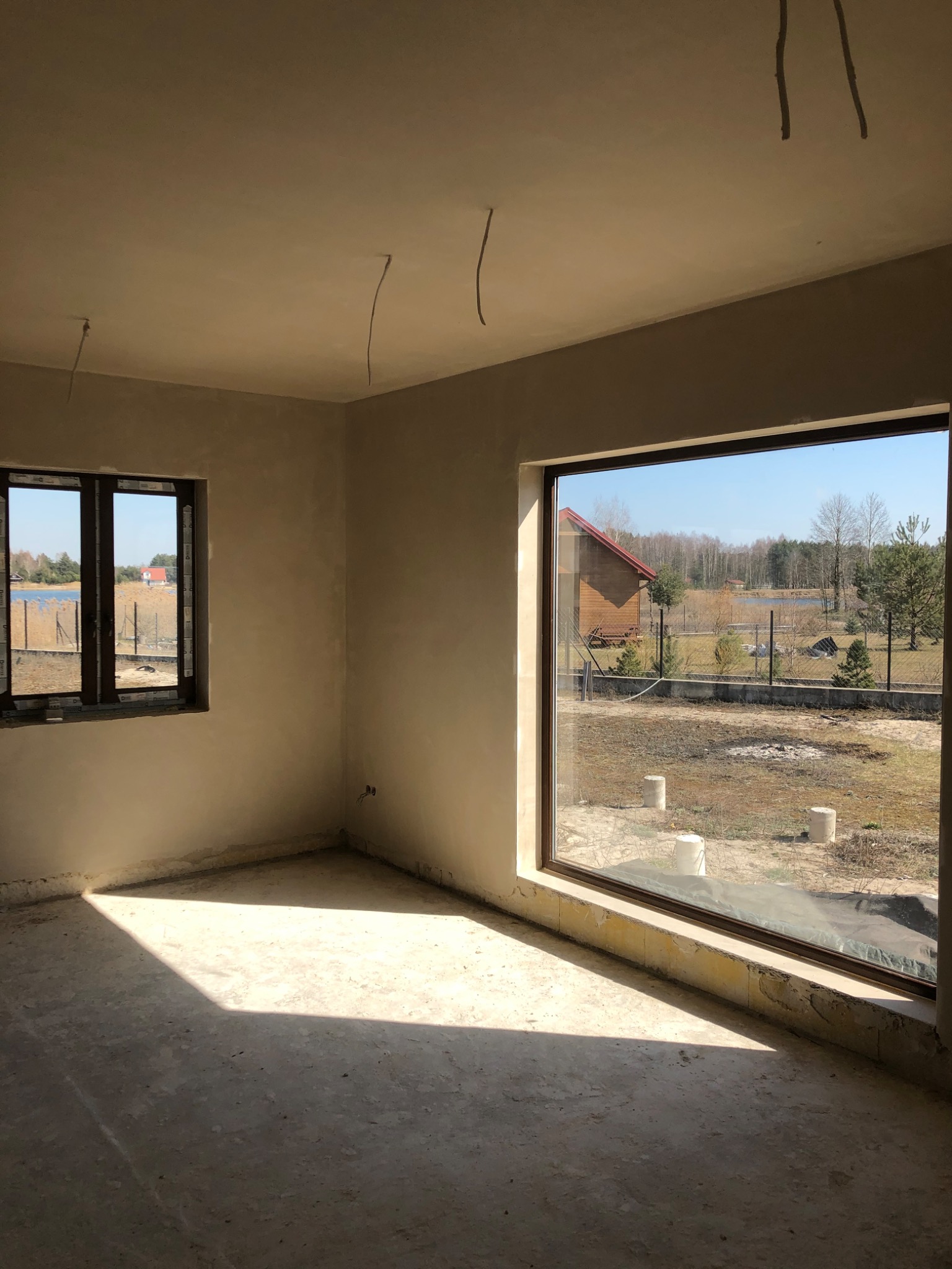 Surowe wnętrze domu w trakcie wykańczania, ściany po wstępnym wyrównaniu, widoczne wystające przewody elektryczne z sufitu, duże okno z widokiem na jezioro i las.