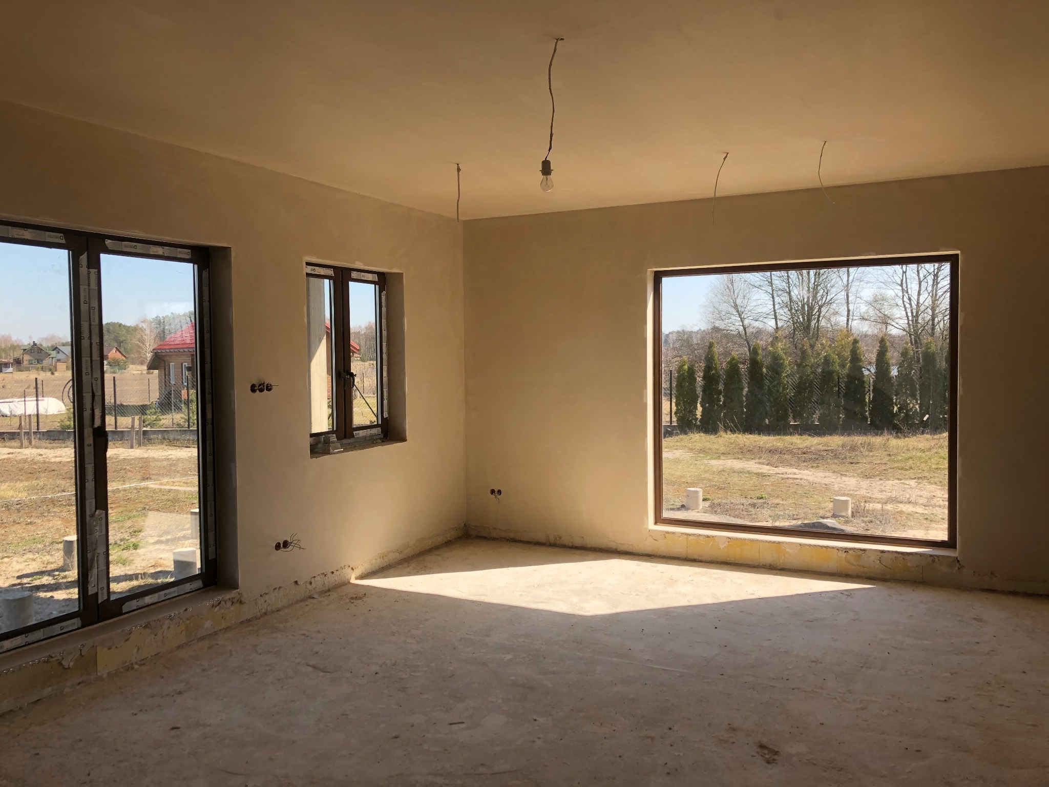 Wnętrze domu w stanie surowym zamkniętym po nałożeniu tynku, widoczne dwa okna z brązowymi ramami i jedno mniejsze, ściany w jasnym odcieniu beżu, podłoga betonowa, na suficie wiszące przewody...