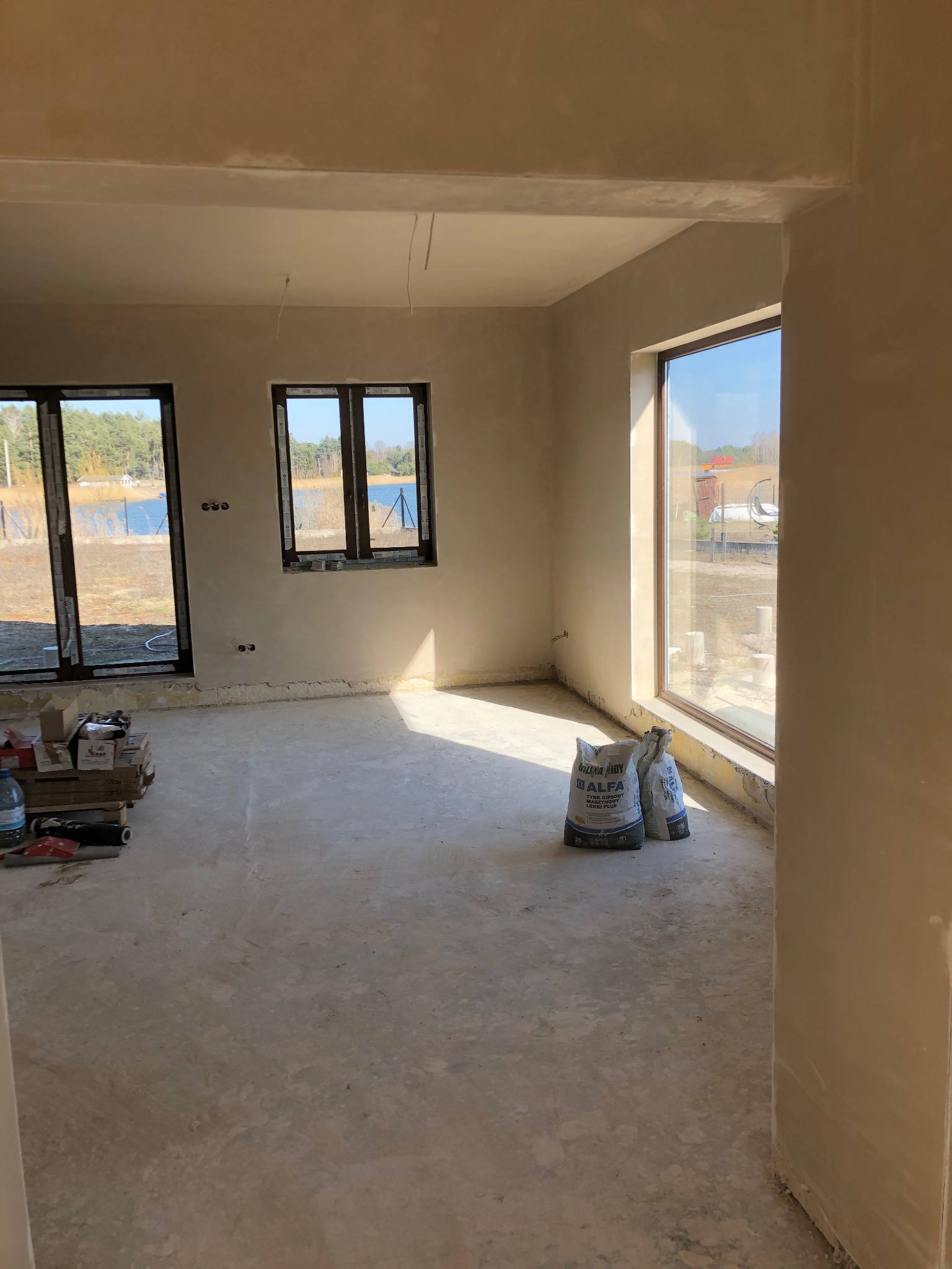 Wnętrze domu w stanie deweloperskim po tynkowaniu maszynowym, widoczne worki z tynkiem gipsowym Alfa Leikki Plus, okna z widokiem na jezioro, podłoga przygotowana do dalszych prac.
