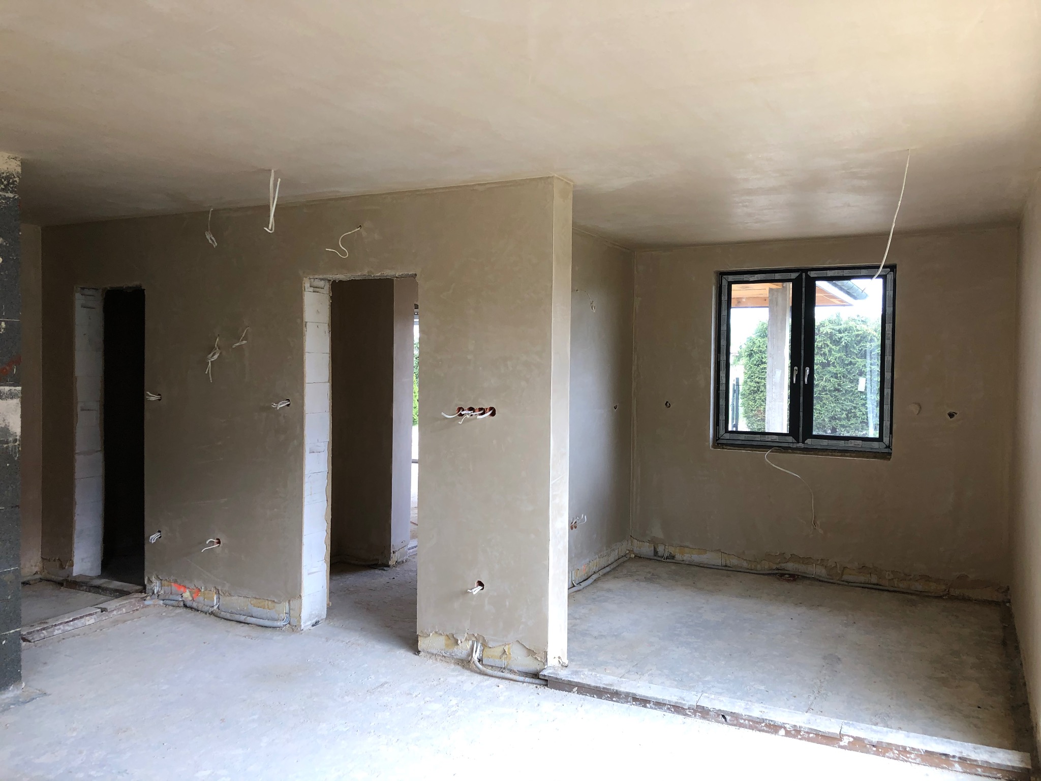 Surowe wnętrze w trakcie remontu: świeżo nałożona warstwa tynku na ścianach i suficie, widoczne wystające przewody elektryczne, przygotowane otwory drzwiowe i okno z widokiem na zieleń.