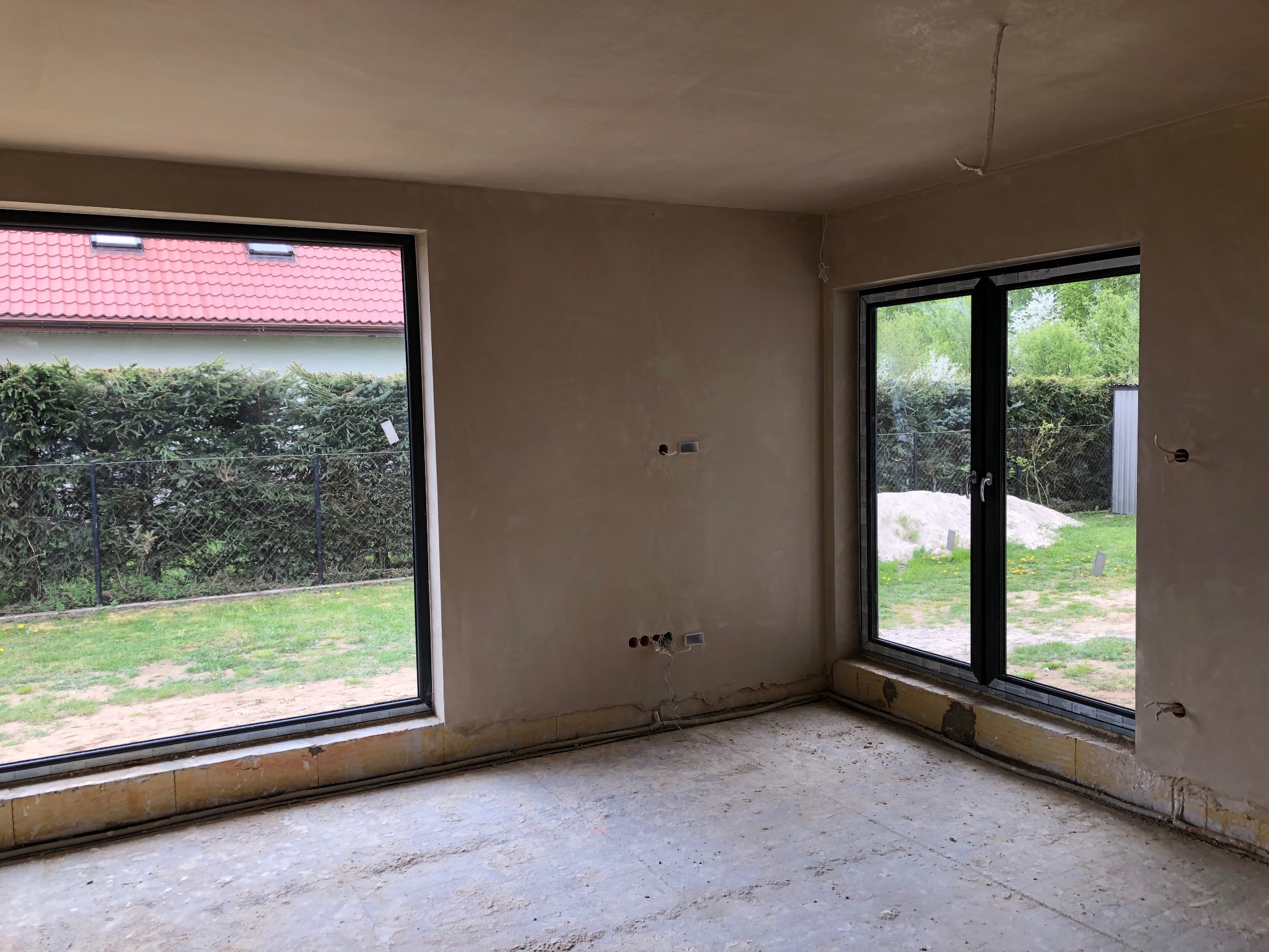 Surowe wnętrze pokoju po nałożeniu tynku, widoczne okna z czarnymi ramami, wystające kable elektryczne i niedokończone wykończenie podłogi.