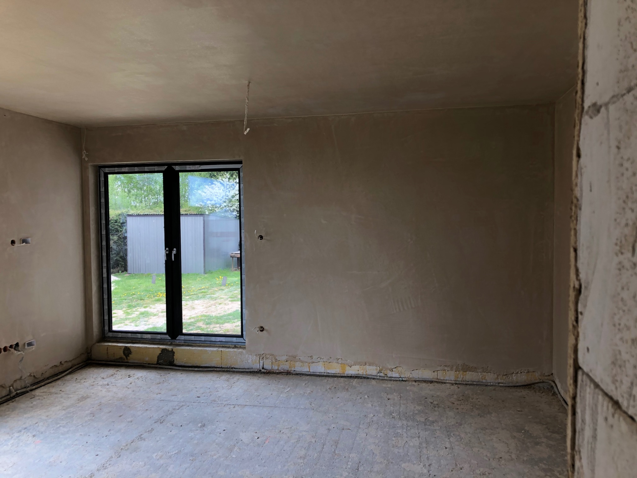 Surowe wnętrze pokoju po nałożeniu wstępnej warstwy tynku, widoczne wystające kable elektryczne i niedoskonałości podłogi, okno z widokiem na ogród z metalowym schowkiem.