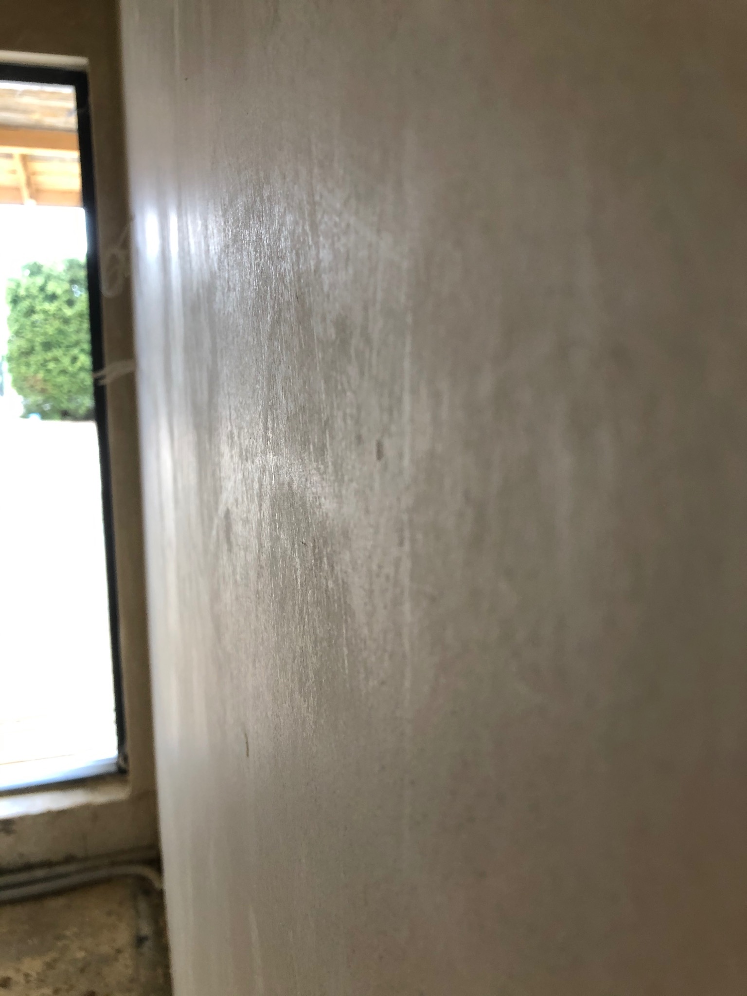 Ściana po nałożeniu gładzi, widoczne delikatne ślady po szpachlowaniu, obok okno z widokiem na zieleń.