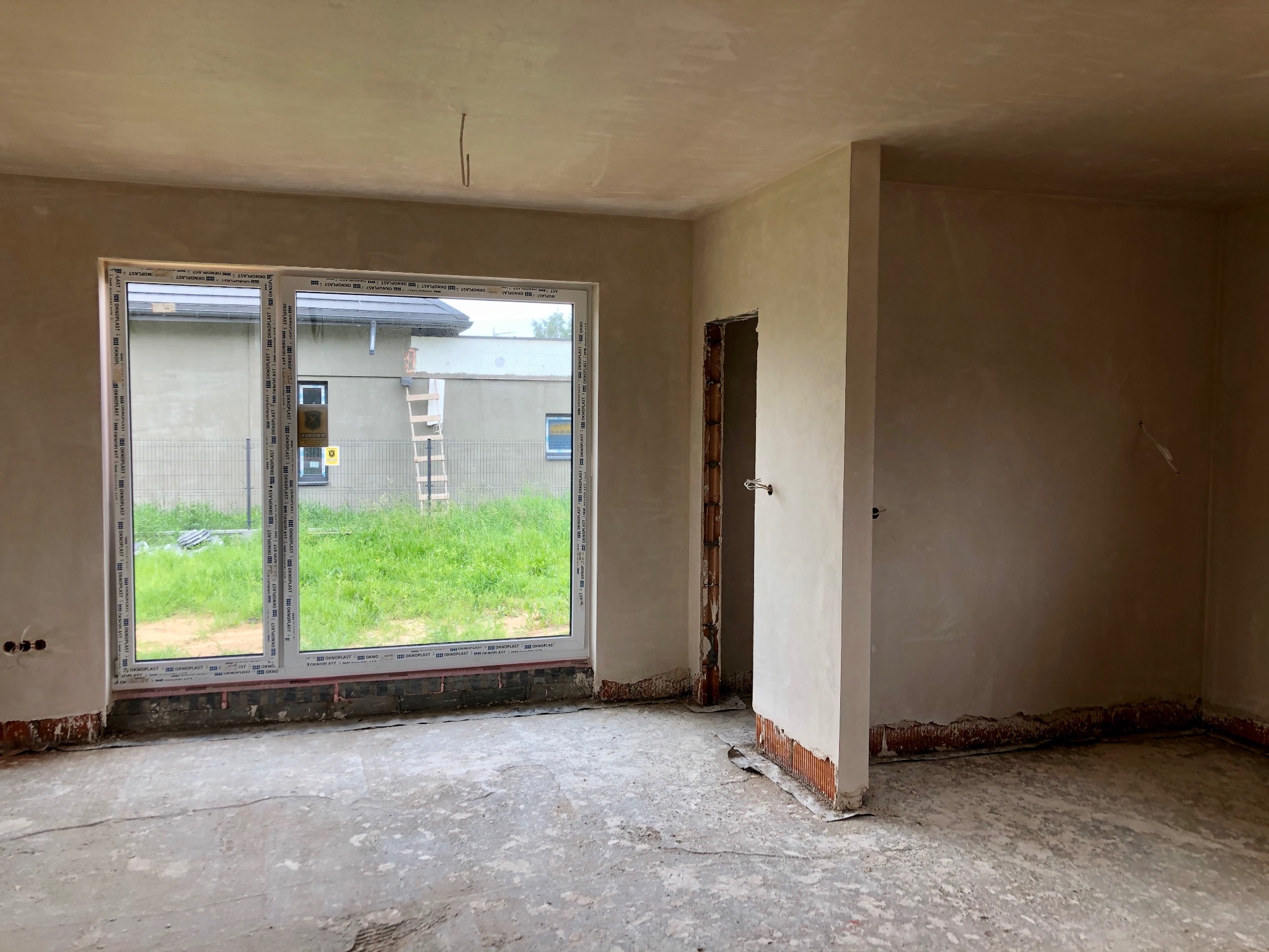 Wnętrze surowego domu po wstępnym wykończeniu ścian, widoczne ślady tynku, duże okno z widokiem na zewnątrz i niedokończone obramowanie drzwi.