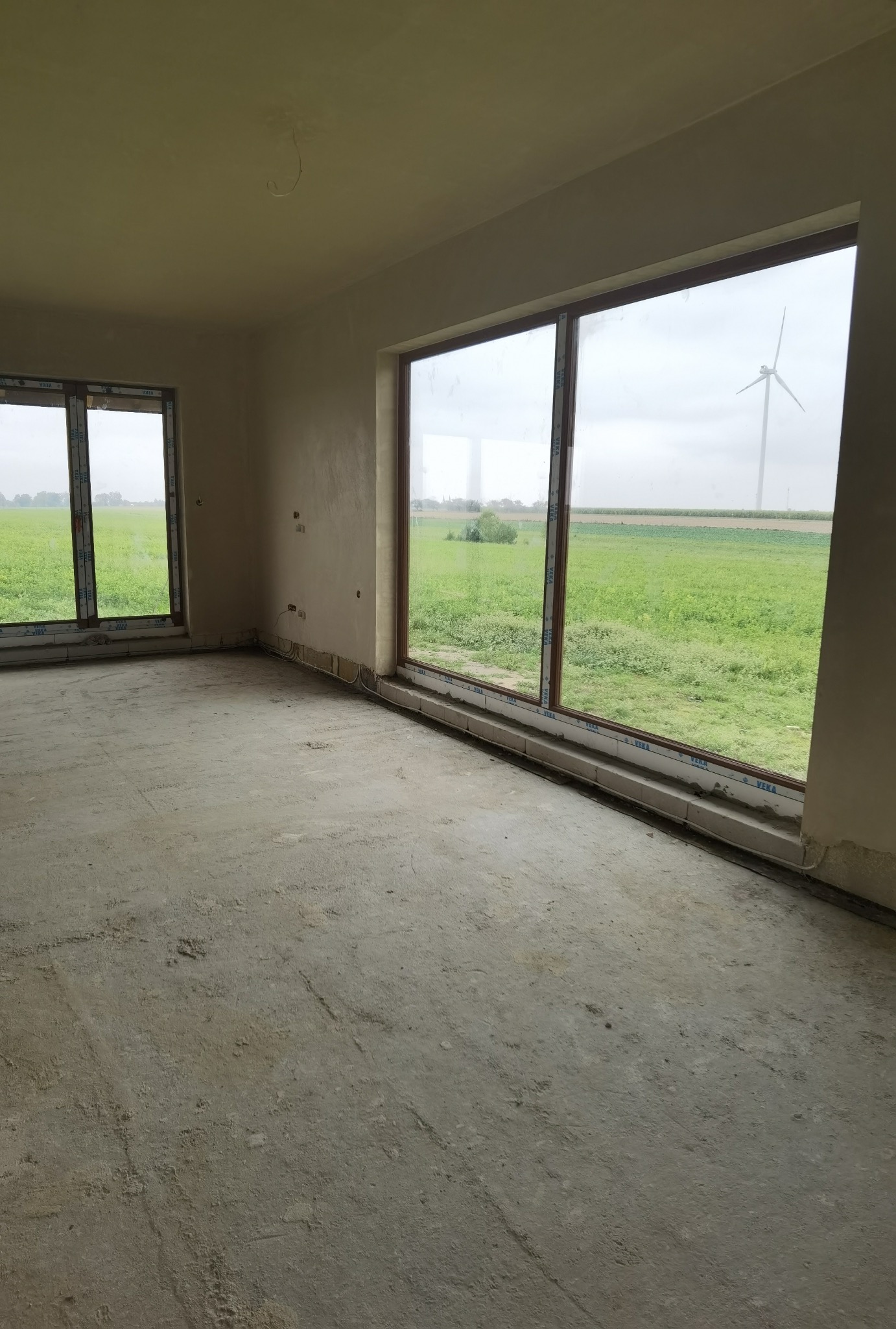 Surowe wnętrze z widokiem na pole i turbinę wiatrową przez okna, ściany przygotowane pod wykończenie, widoczne ślady po pracach budowlanych na podłodze.