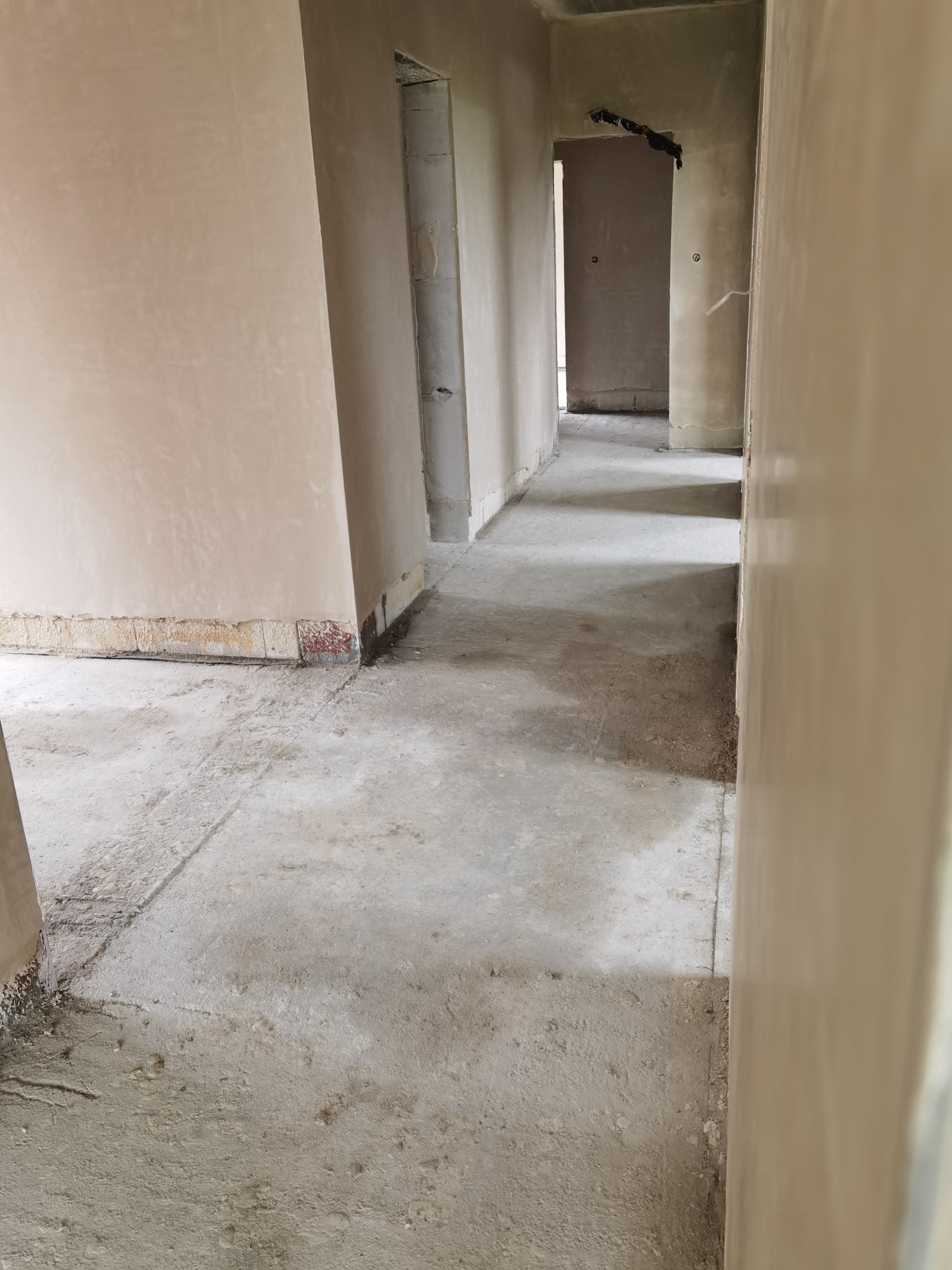 Wnętrze w trakcie prac wykończeniowych: ściany pokryte jasnym tynkiem, surowy beton na podłodze, widoczne fragmenty instalacji elektrycznej wystające ze ściany, kilka otworów drzwiowych...