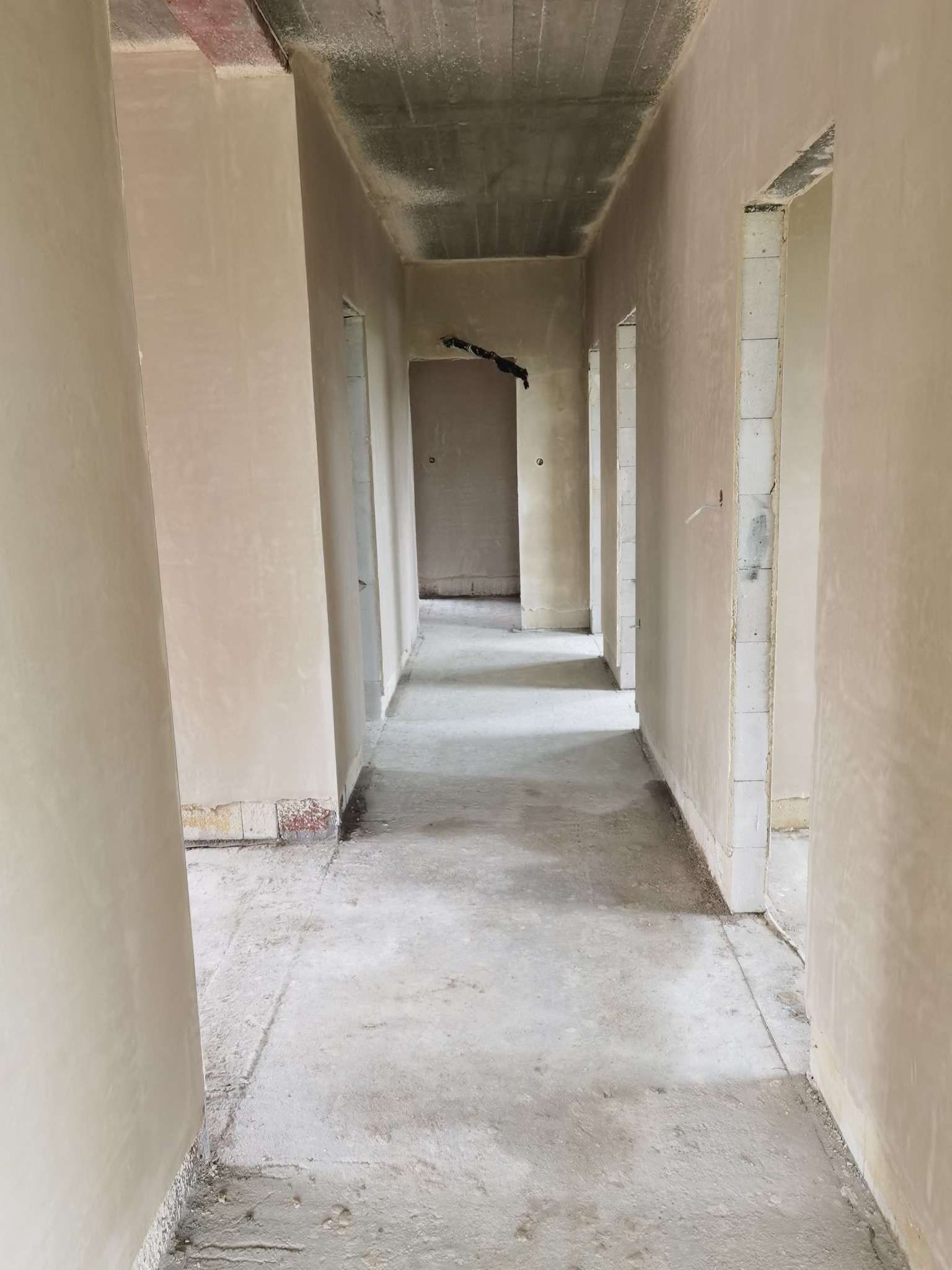 Korytarz w surowym stanie po tynkowaniu maszynowym, widoczne ślady zaprawy na podłodze, brak wykończenia, surowy betonowy strop.