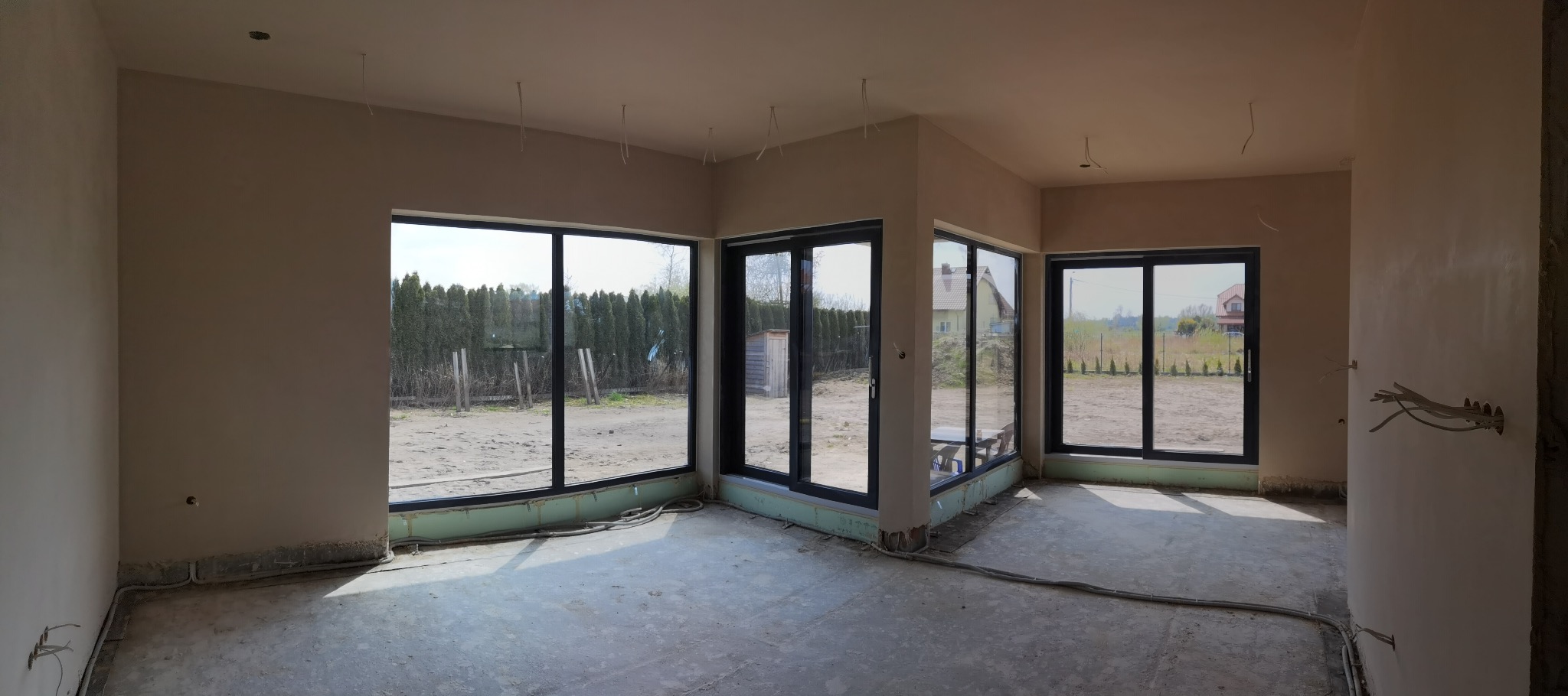 Surowe wnętrze domu w trakcie wykańczania, ściany i sufit po wstępnym wygładzeniu, widoczne kable elektryczne, duże okna z widokiem na ogród.