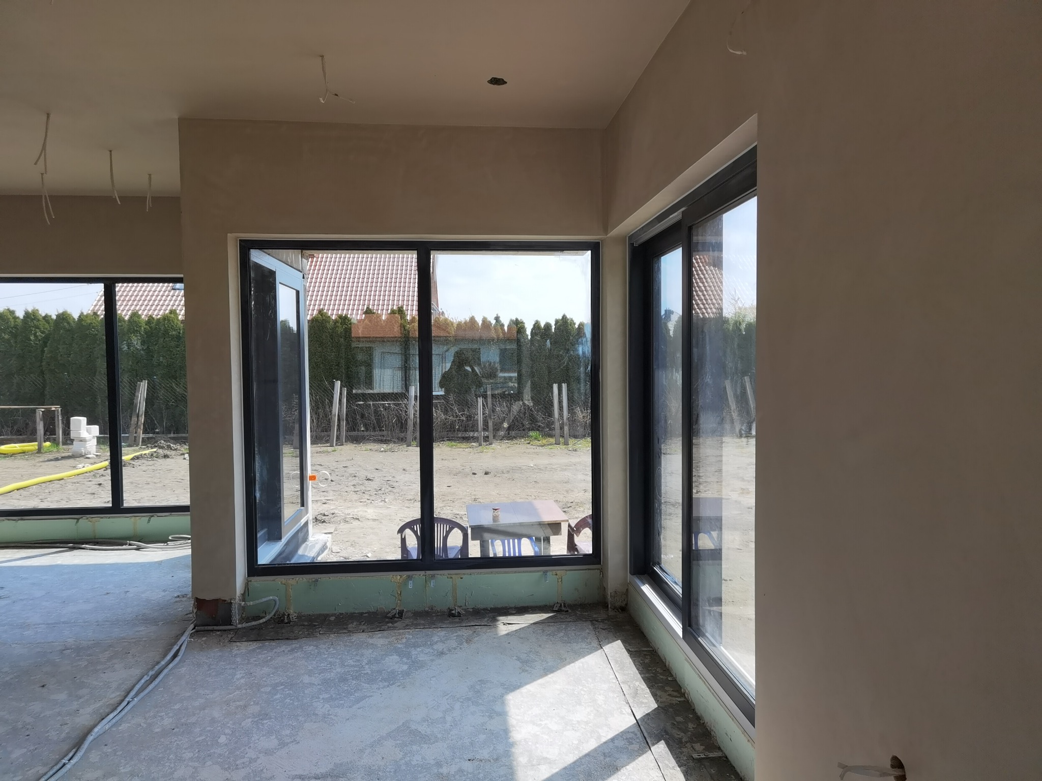Surowe ściany w nowym domu z dużymi oknami, widoczne przewody elektryczne wystające ze ścian, podłoga w stanie surowym, widok na ogród przez okna.