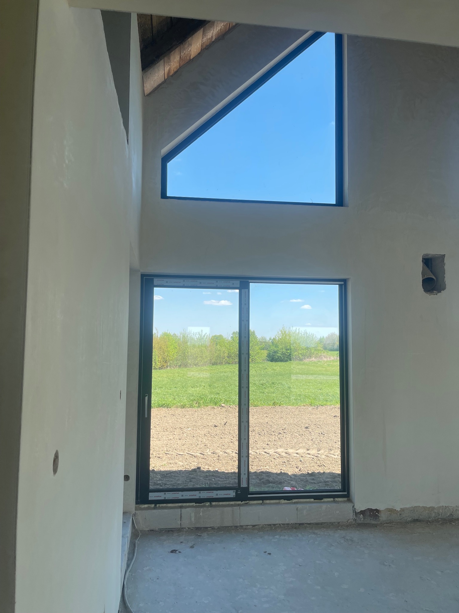 Wnętrze w stanie deweloperskim z oknami na zewnątrz, ściany po nałożeniu gładzi, widoczne niedoskonałości podłoża i brak wykończenia podłogi.