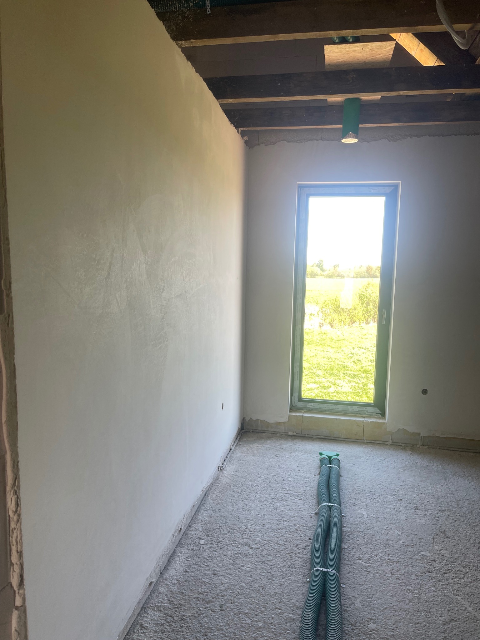 Świeżo otynkowana ściana w surowym wnętrzu z widokiem na zewnątrz przez okno; na podłodze leżą zielone rury wentylacyjne.