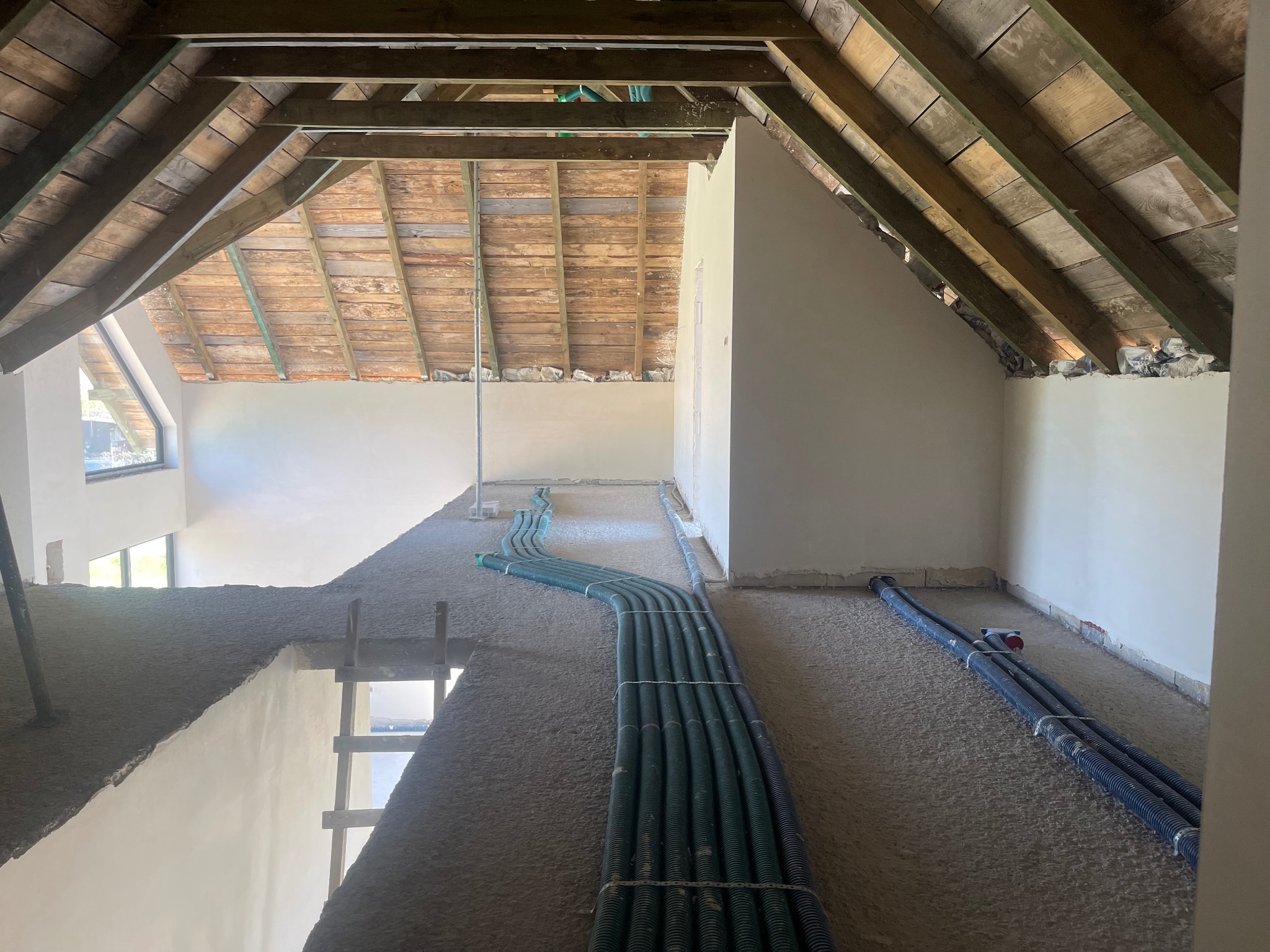 Poddasze w trakcie prac wykończeniowych, widoczne surowe drewniane krokwie, ściany pokryte szarą warstwą, prawdopodobnie tynkiem, oraz rury instalacyjne ułożone na podłodze.