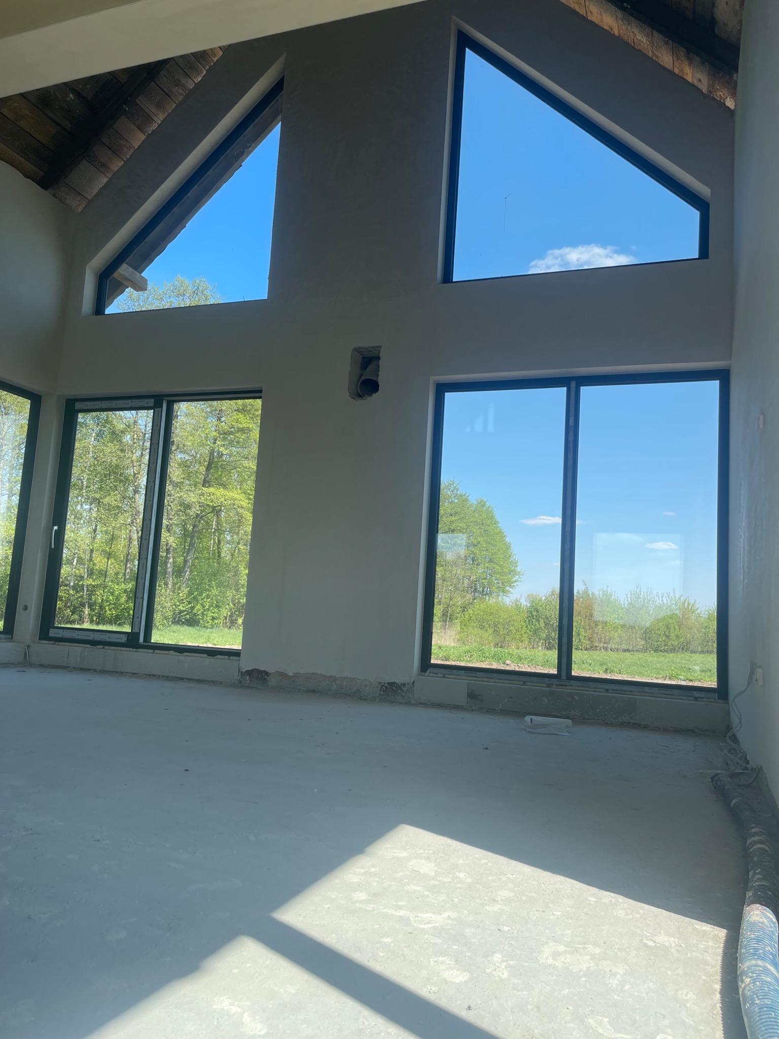 Surowe wnętrze domu z dużymi oknami, widoczne ślady po pracach budowlanych na ścianach i podłodze, przygotowane do dalszego wykończenia.