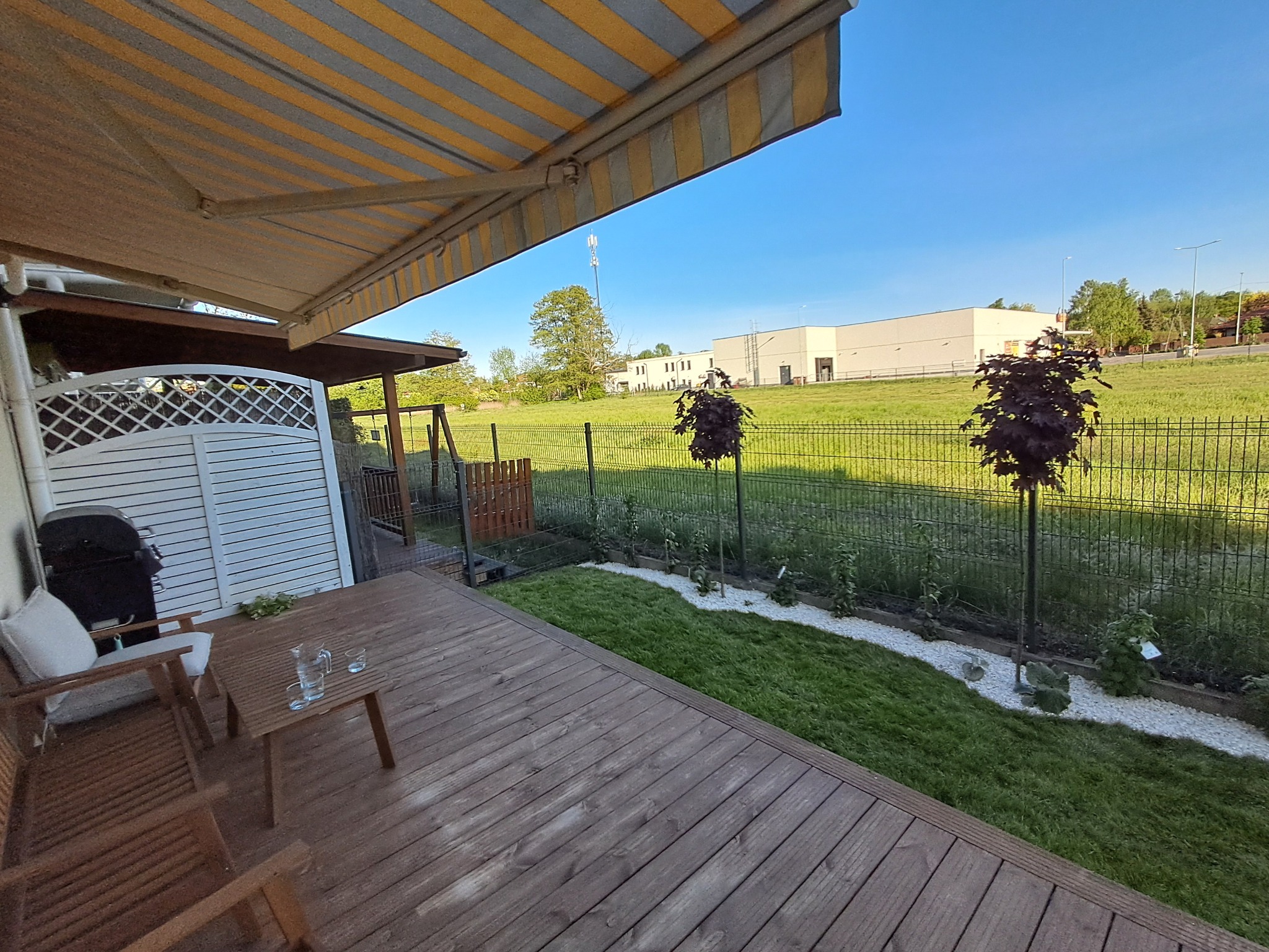 Ujęcie z tarasu na ogród z dwoma kulistymi drzewami, ścieżką z białego żwiru i zielonym trawnikiem. W tle budynek i błękitne niebo.