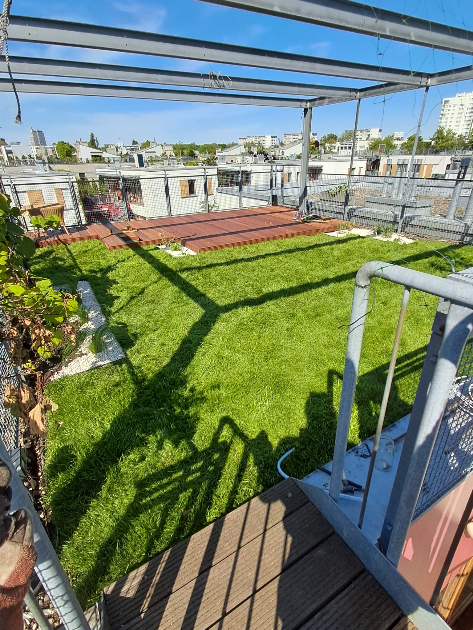 Zadbany trawnik na dachu budynku, otoczony drewnianym tarasem i białymi kamykami. Metalowa konstrukcja nad ogrodem. Widok z góry na zieloną oazę na wysokości.