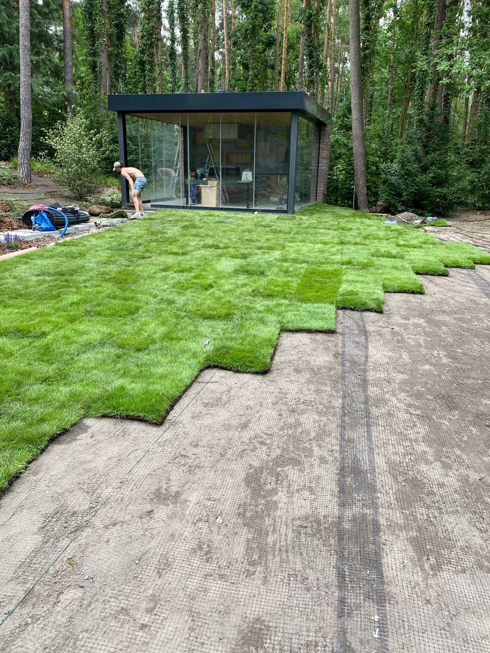 Układanie trawy z rolki na przygotowanym podłożu w ogrodzie z nowoczesną altaną. Widoczny pracownik w trakcie pracy i częściowo ułożona trawa.