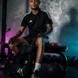 NowakFoto (Calor Sp. z o.o.) - Mężczyzna w czarnej koszulce z logo Adidas siedzi na ławce w siłowni, obok torba sportowa, w tle hantle i sztanga, oświetlenie neonowe.