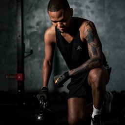 NowakFoto (Calor Sp. z o.o.) - Mężczyzna w czarnej koszulce na ramiączkach i spodenkach klęczy na jednym kolanie, trzymając kettlebell w rękawicy z logo Nike, z tatuażem na ramieniu i złotą bransoletką na nadgarstku, na tle...