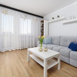 NowakFoto (Calor Sp. z o.o.) - Jasne wnętrze salonu z szarą sofą, białym stolikiem kawowym z żółtymi tulipanami w wazonie, drewnianą podłogą i oknem z białymi zasłonami ozdobionym gwiazdami.