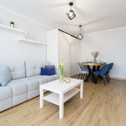 NowakFoto (Calor Sp. z o.o.) - Jasne, minimalistyczne wnętrze salonu z sofą, stolikiem kawowym z tulipanami w wazonie, okrągłym stołem jadalnym i dekoracyjnymi półkami na ścianie.