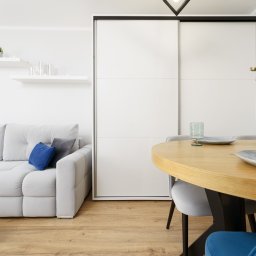 NowakFoto (Calor Sp. z o.o.) - Jasne wnętrze: szara sofa z niebieskimi poduszkami, okrągły drewniany stół z nakryciem i wazonem z różowymi goździkami, biała szafa przesuwna w tle.