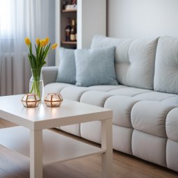 NowakFoto (Calor Sp. z o.o.) - Jasny salon z białym narożnikiem, stolikiem kawowym z żółtymi tulipanami w wazonie i dwiema świecami w szklanych lampionach.