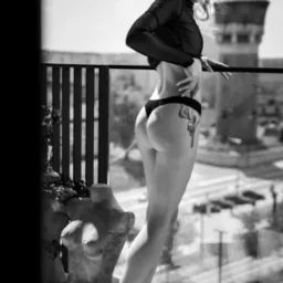 Czarno-białe zdjęcie kobiety w bieliźnie i prześwitującej bluzce, stojącej na balkonie z widokiem na miasto i wieżę ciśnień, z rzeźbą popiersia obok niej i tatuażem na biodrze.