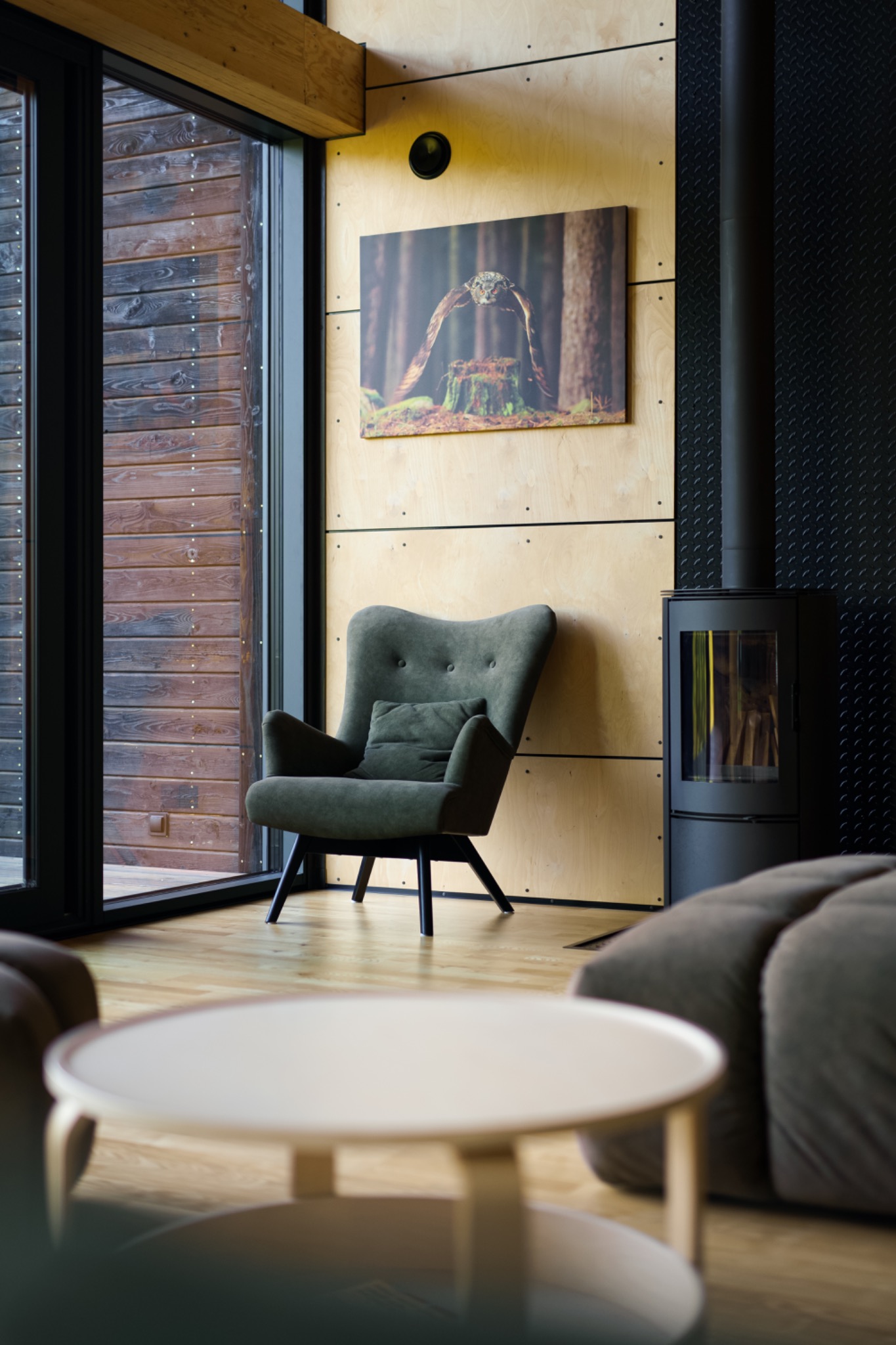 Wnętrze pokoju w Sosnowcu z drewnianymi panelami, ciemnozielonym fotelem z poduszką, czarnym kominkiem i obrazem sowy na ścianie.