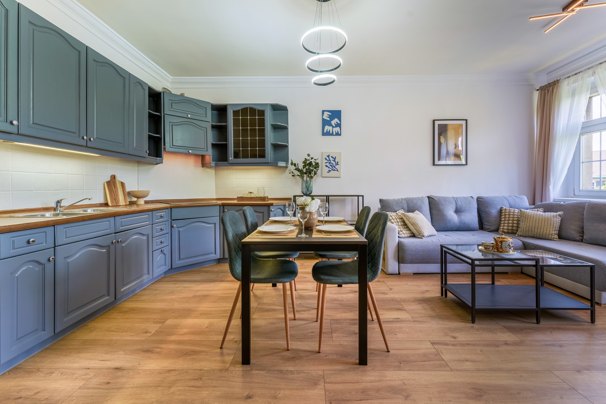 Jasne wnętrze mieszkania z szarymi szafkami kuchennymi, drewnianym blatem, jadalnią z czarnym stołem i zielonymi krzesłami oraz szarą sofą narożną w tle.