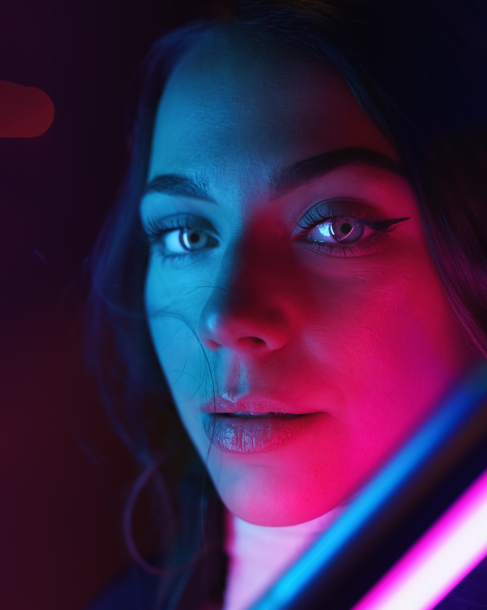 Portret kobiety z makijażem oczu, oświetlonej neonowym światłem w odcieniach różu i błękitu, z widoczną częścią neonowej lampy w kadrze.