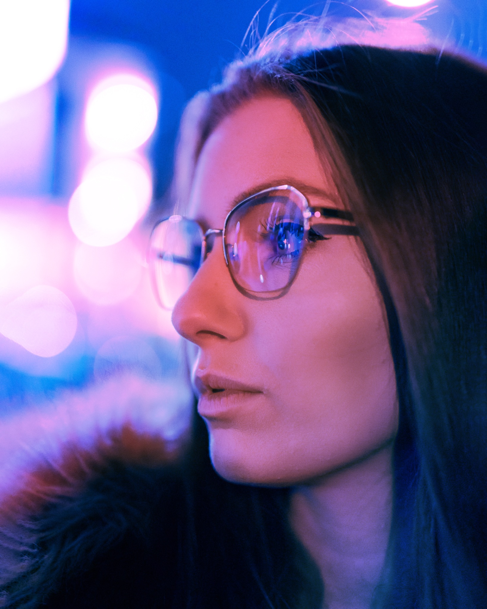 Portret kobiety w okularach z odbiciem w szkłach, oświetlonej neonowym, niebiesko-różowym światłem.