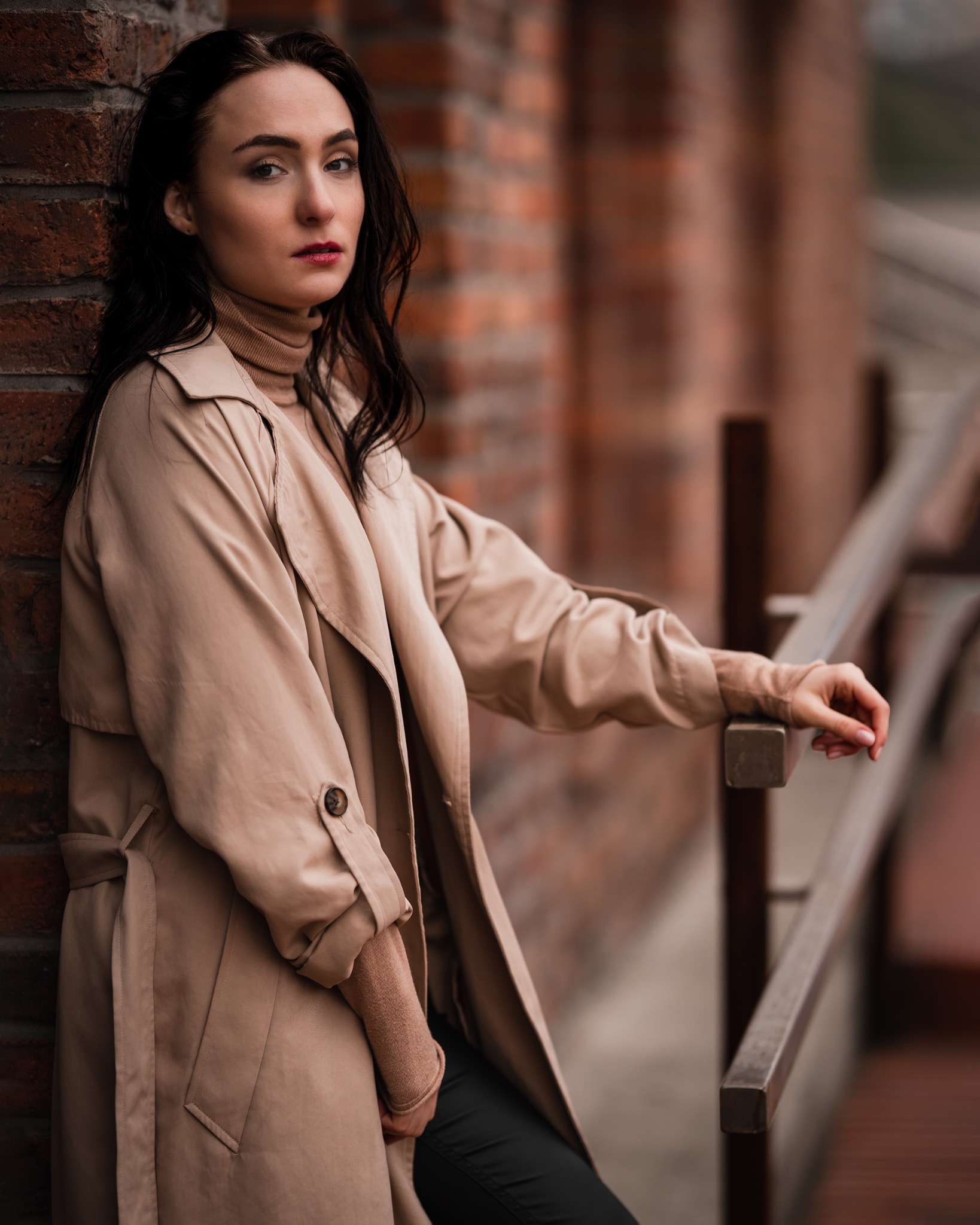 Portret kobiety w beżowym płaszczu, opierającej się o ceglaną ścianę i drewnianą barierkę na zewnątrz.