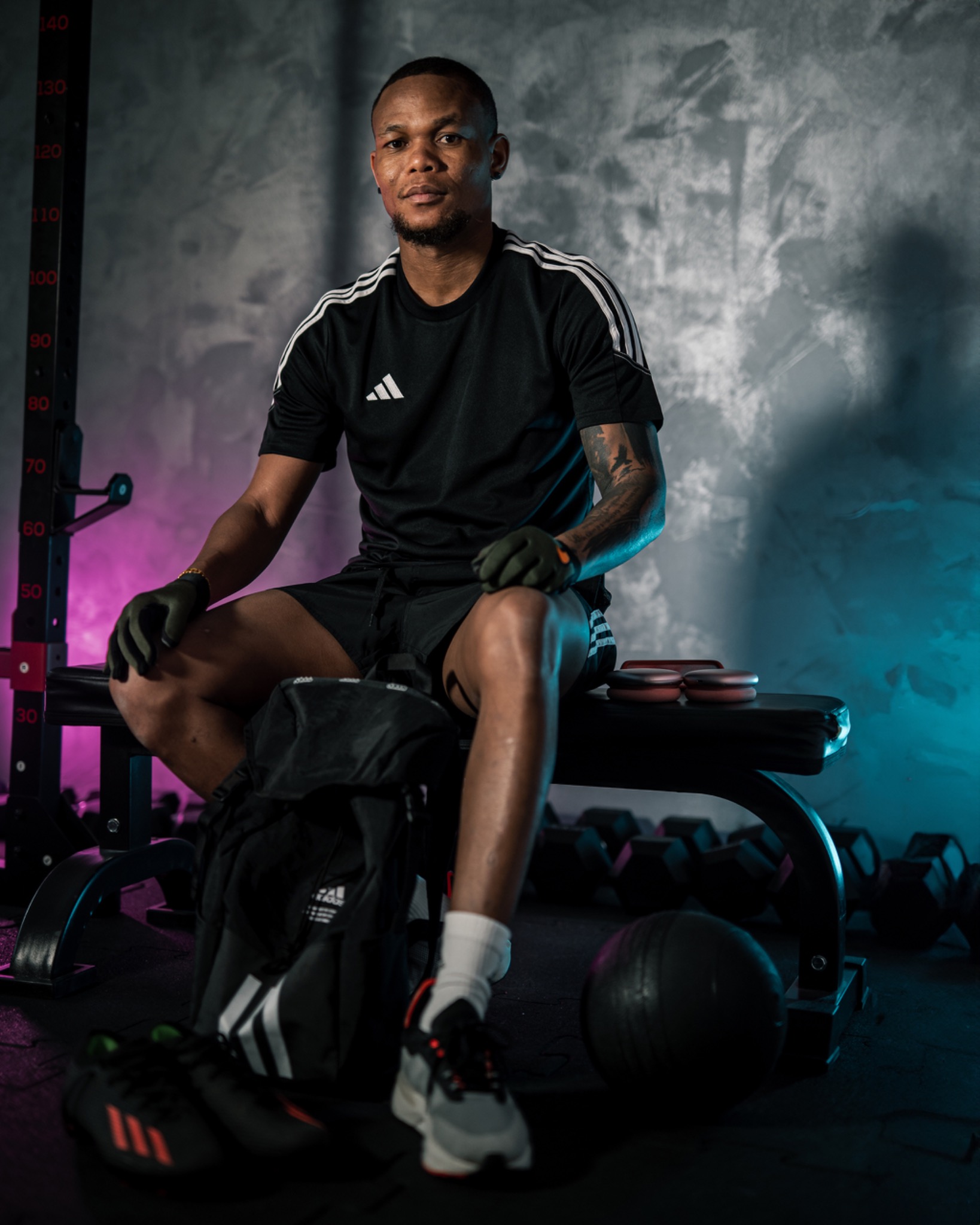 Mężczyzna w czarnej koszulce z logo Adidas siedzi na ławce w siłowni, obok torba sportowa, w tle hantle i sztanga, oświetlenie neonowe.