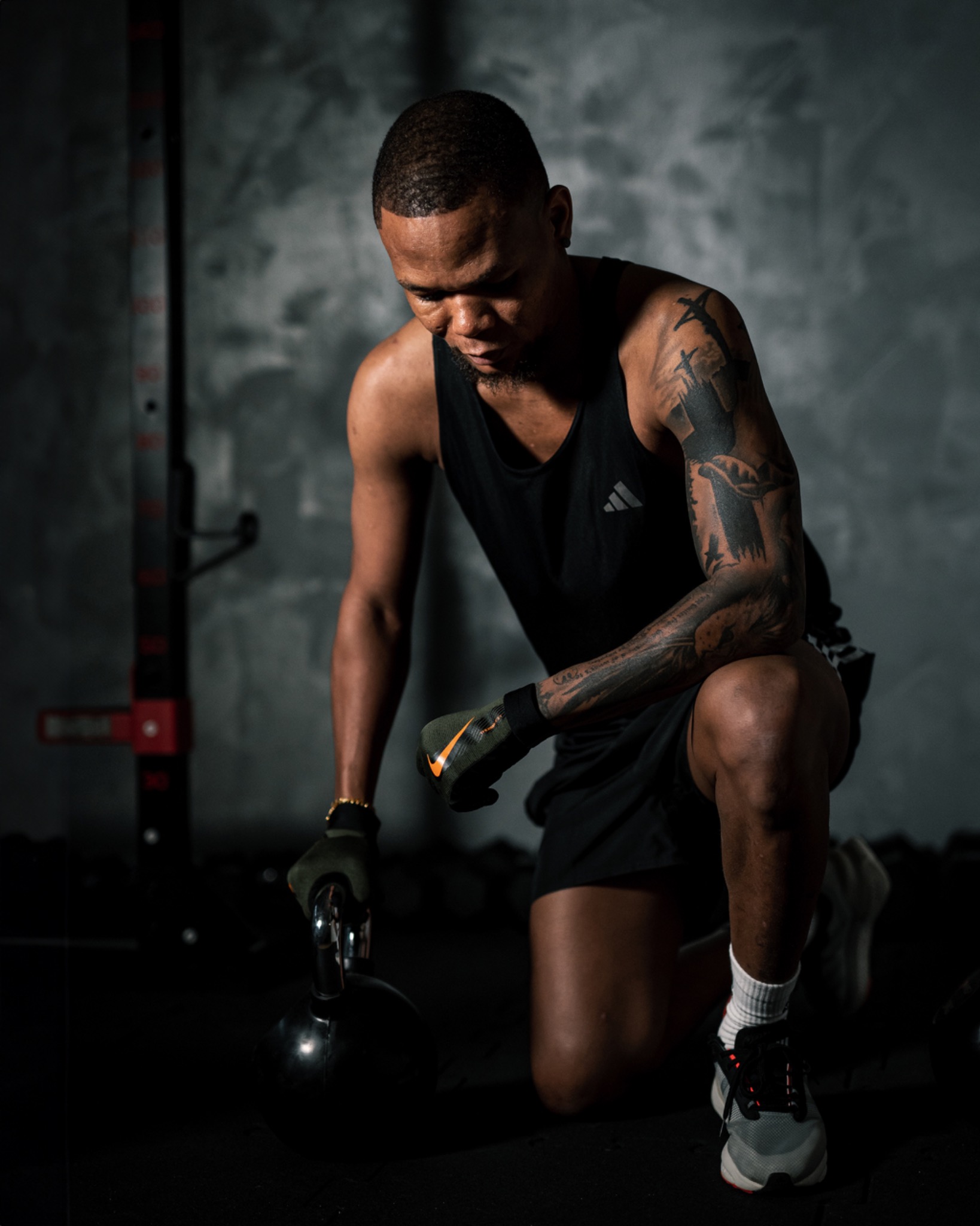 Mężczyzna w czarnej koszulce na ramiączkach i spodenkach klęczy na jednym kolanie, trzymając kettlebell w rękawicy z logo Nike, z tatuażem na ramieniu i złotą bransoletką na nadgarstku, na tle...
