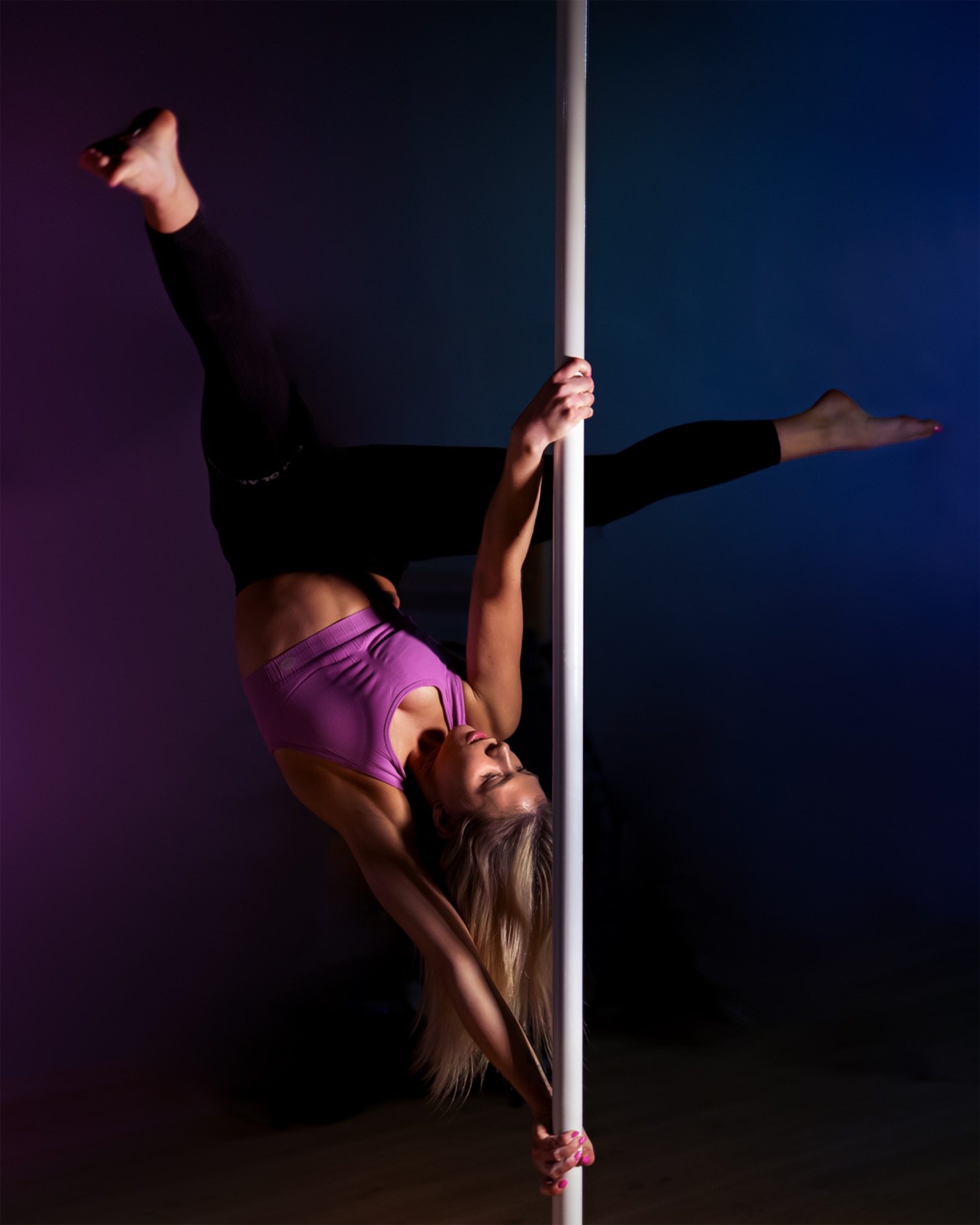 Kobieta w sportowym stroju, wykonująca akrobacje na rurze, z nogami uniesionymi do góry, w dynamicznej pozie na tle ciemnego studia z kolorowym oświetleniem.