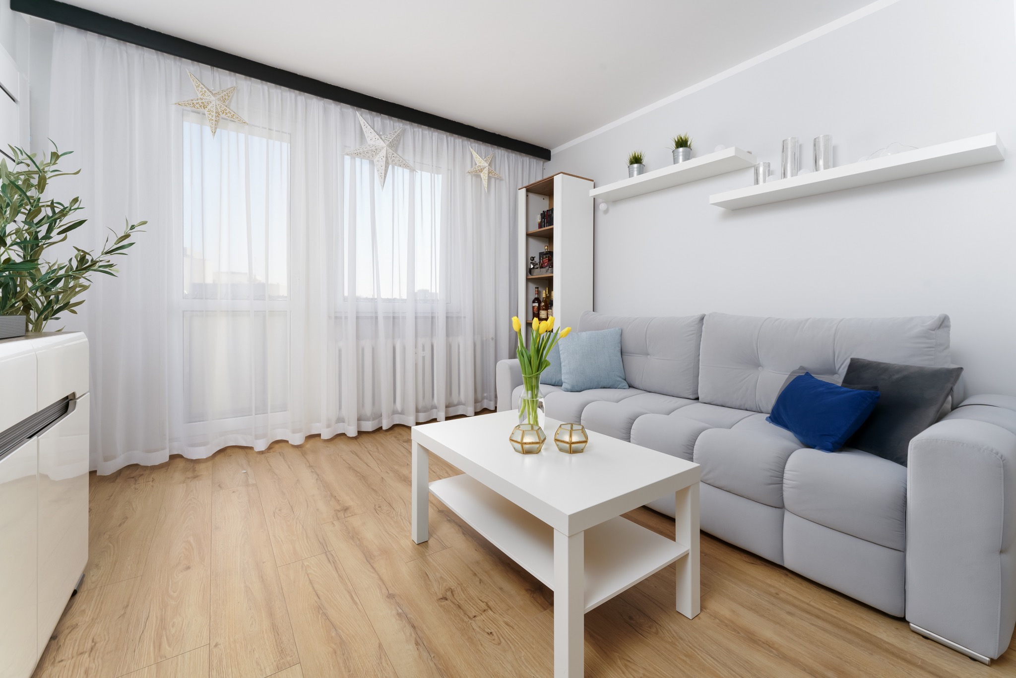 Jasne wnętrze salonu z szarą sofą, białym stolikiem kawowym z żółtymi tulipanami w wazonie, drewnianą podłogą i oknem z białymi zasłonami ozdobionym gwiazdami.