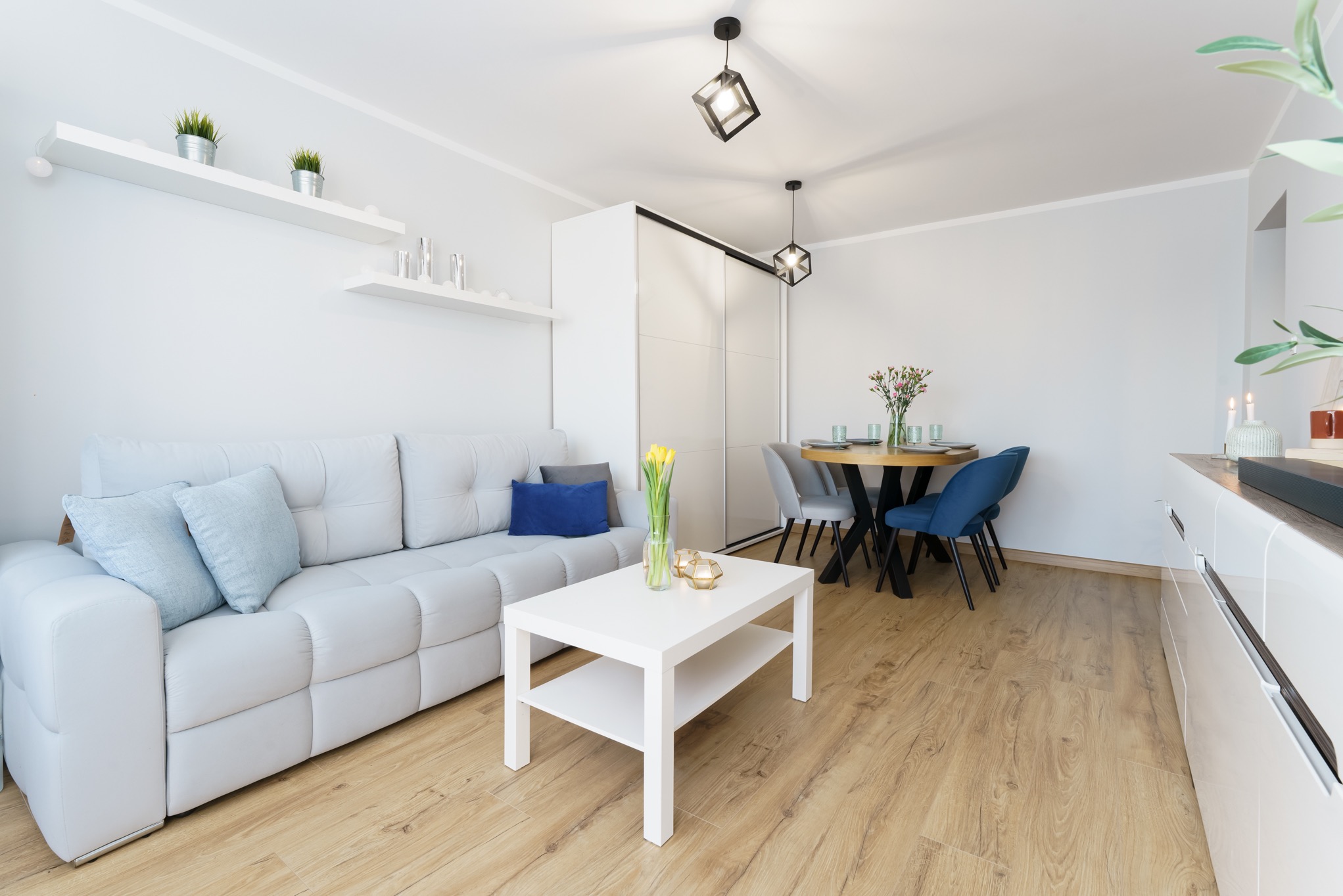 Jasne, minimalistyczne wnętrze salonu z sofą, stolikiem kawowym z tulipanami w wazonie, okrągłym stołem jadalnym i dekoracyjnymi półkami na ścianie.