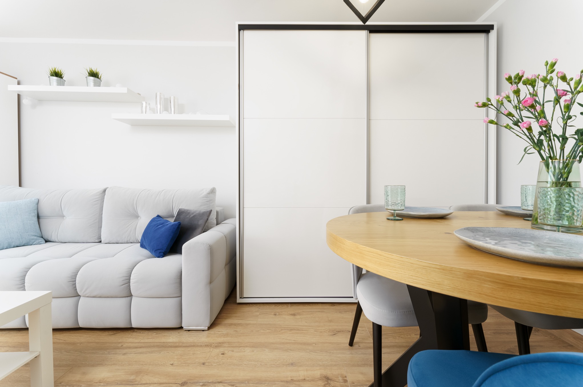 Jasne wnętrze: szara sofa z niebieskimi poduszkami, okrągły drewniany stół z nakryciem i wazonem z różowymi goździkami, biała szafa przesuwna w tle.