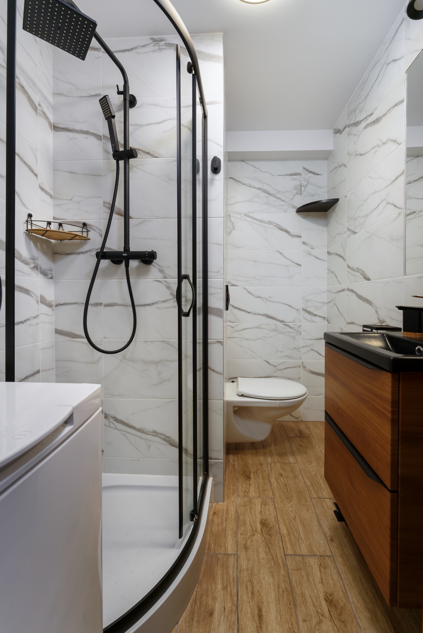 Nowoczesna łazienka z kabiną prysznicową typu walk-in z czarnymi ramami, białymi płytkami imitującymi marmur, drewnianą szafką pod umywalką i drewnopodobną podłogą.