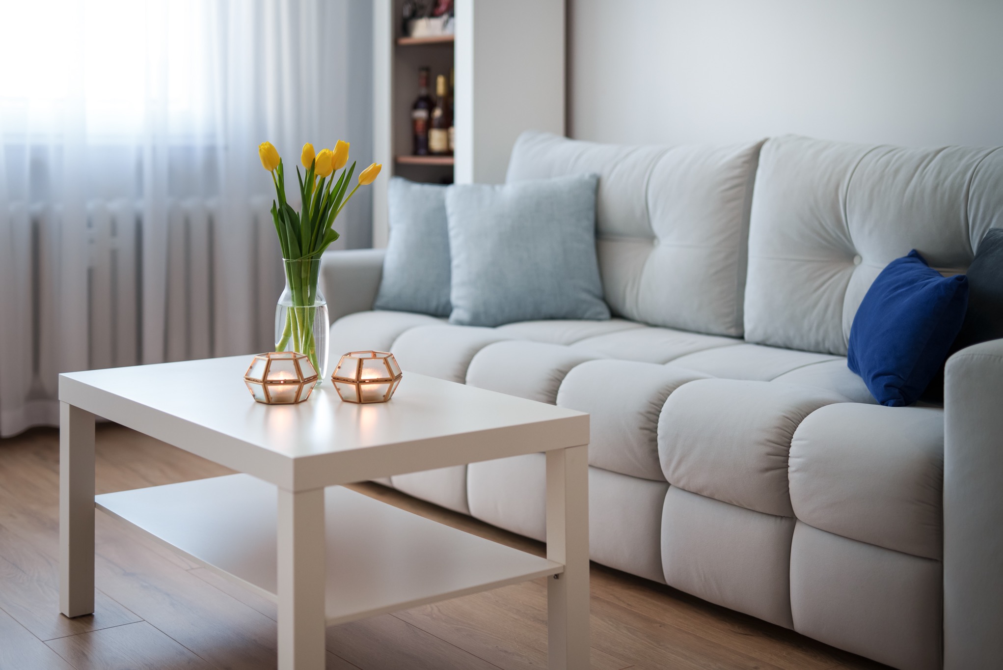 Jasny salon z białym narożnikiem, stolikiem kawowym z żółtymi tulipanami w wazonie i dwiema świecami w szklanych lampionach.
