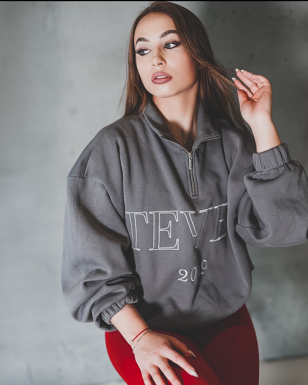 Portret kobiety w szarej bluzie z napisem, z delikatnym makijażem i uniesioną dłonią we włosach, na tle betonowej ściany.