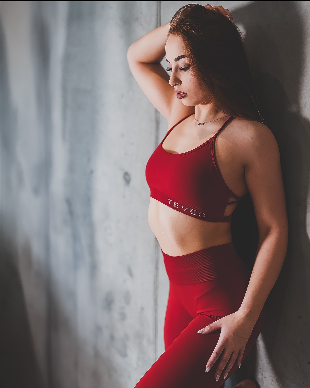 Kobieta w czerwonym sportowym stroju TEVEO pozuje oparta o betonową ścianę, z ręką na głowie i drugą na udzie, w subtelnym oświetleniu studyjnym.